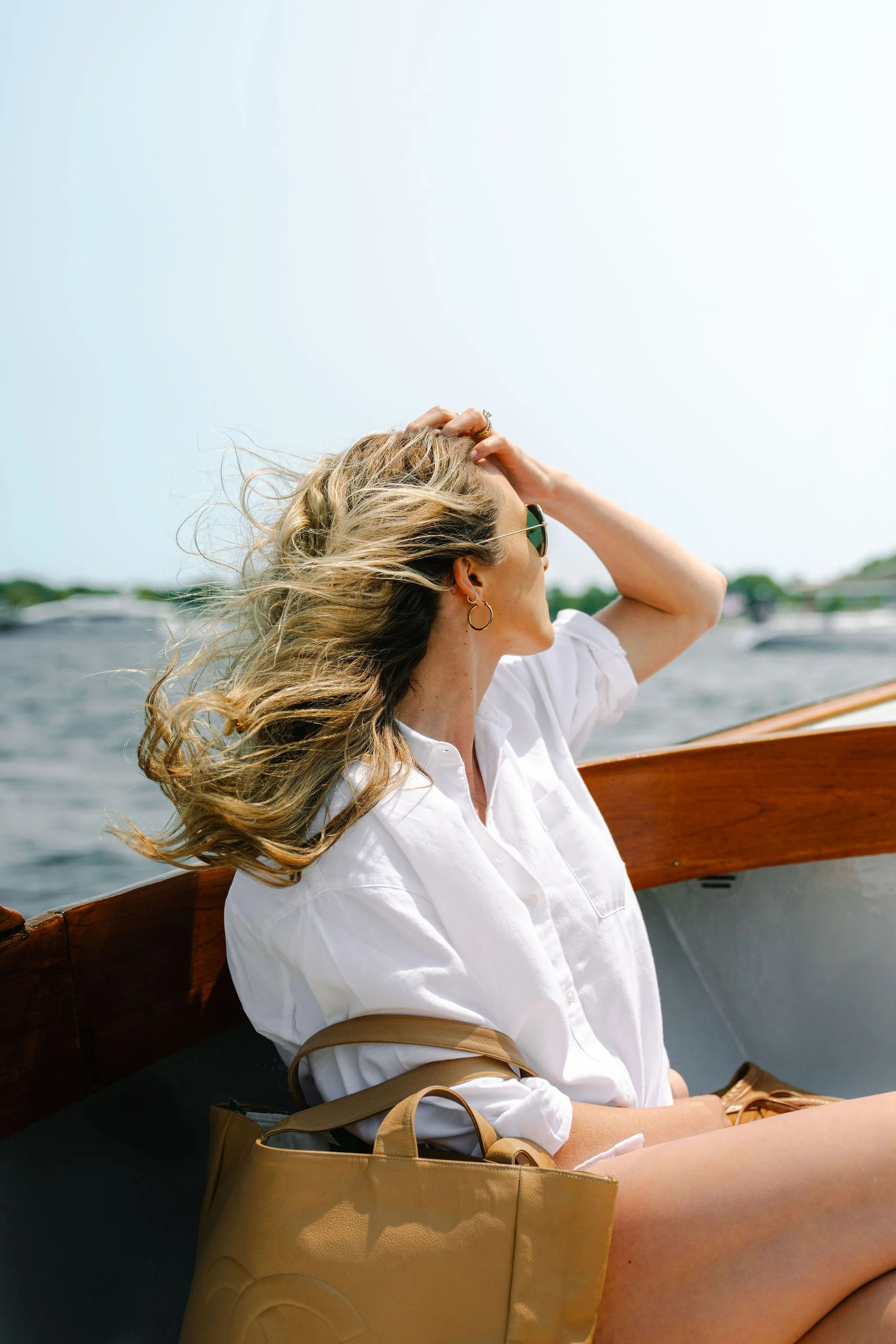 A woman with blonde hair wearing sunglasses and gold hoop earrings sitting on a boat, with water and boats in the background.
