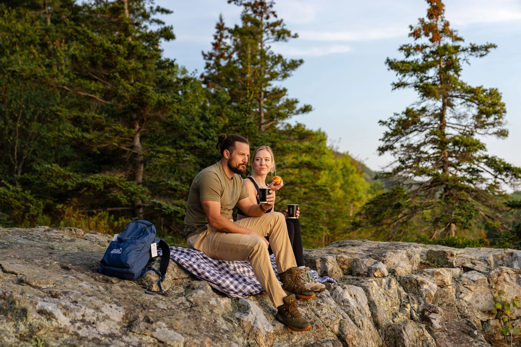 A man and a woman sitting on a rock in a forested area, enjoying drinks and a snack, with a backpack and a blanket nearby.