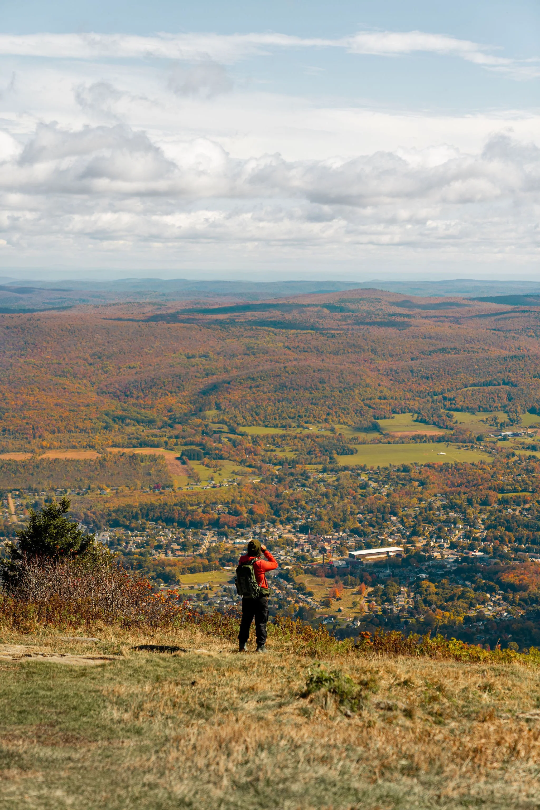 A person standing on a grassy hillside with a backpack, looking out over a town and rolling hills covered in autumn foliage.