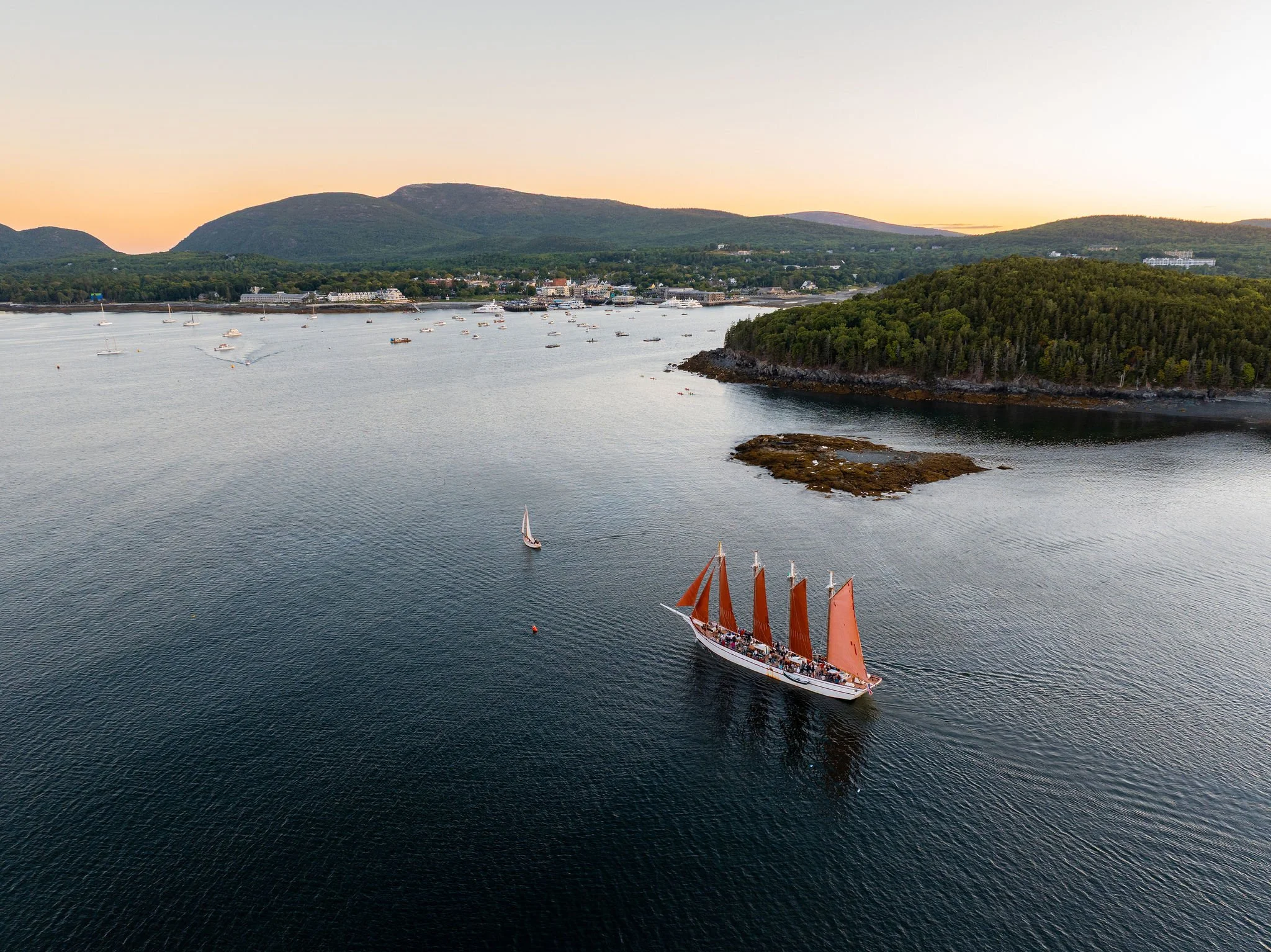A sailboat with orange sails sailing on calm water near a small forested island, with a coastal town and mountains in the background during sunset.