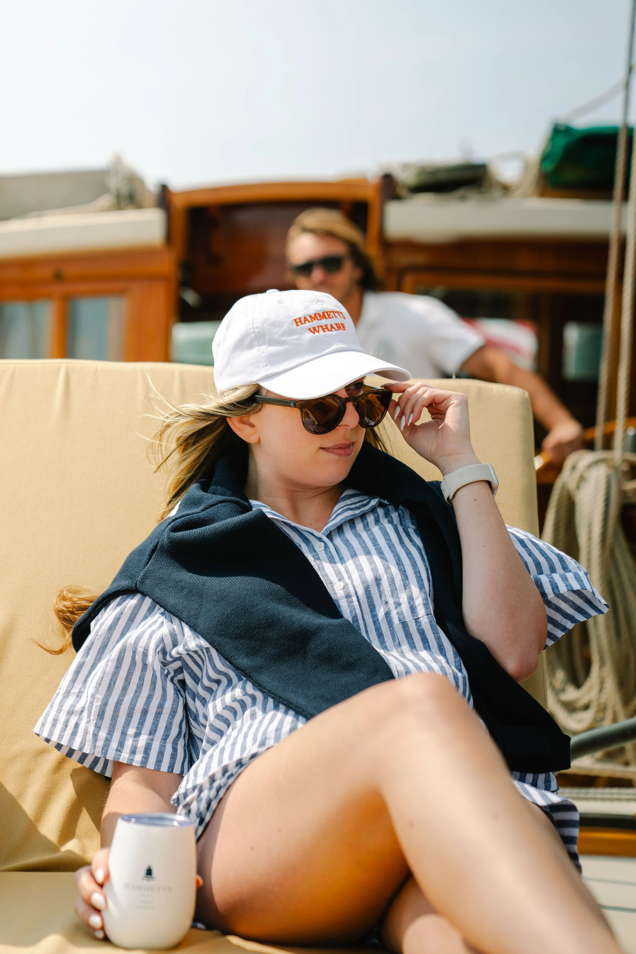 A woman sitting on a beige outdoor lounge chair, wearing sunglasses, a white cap, a blue striped shirt, and a black sweater draped over her shoulders. She's holding a white cup and sitting next to a boat, with a man in the background in front of a wo