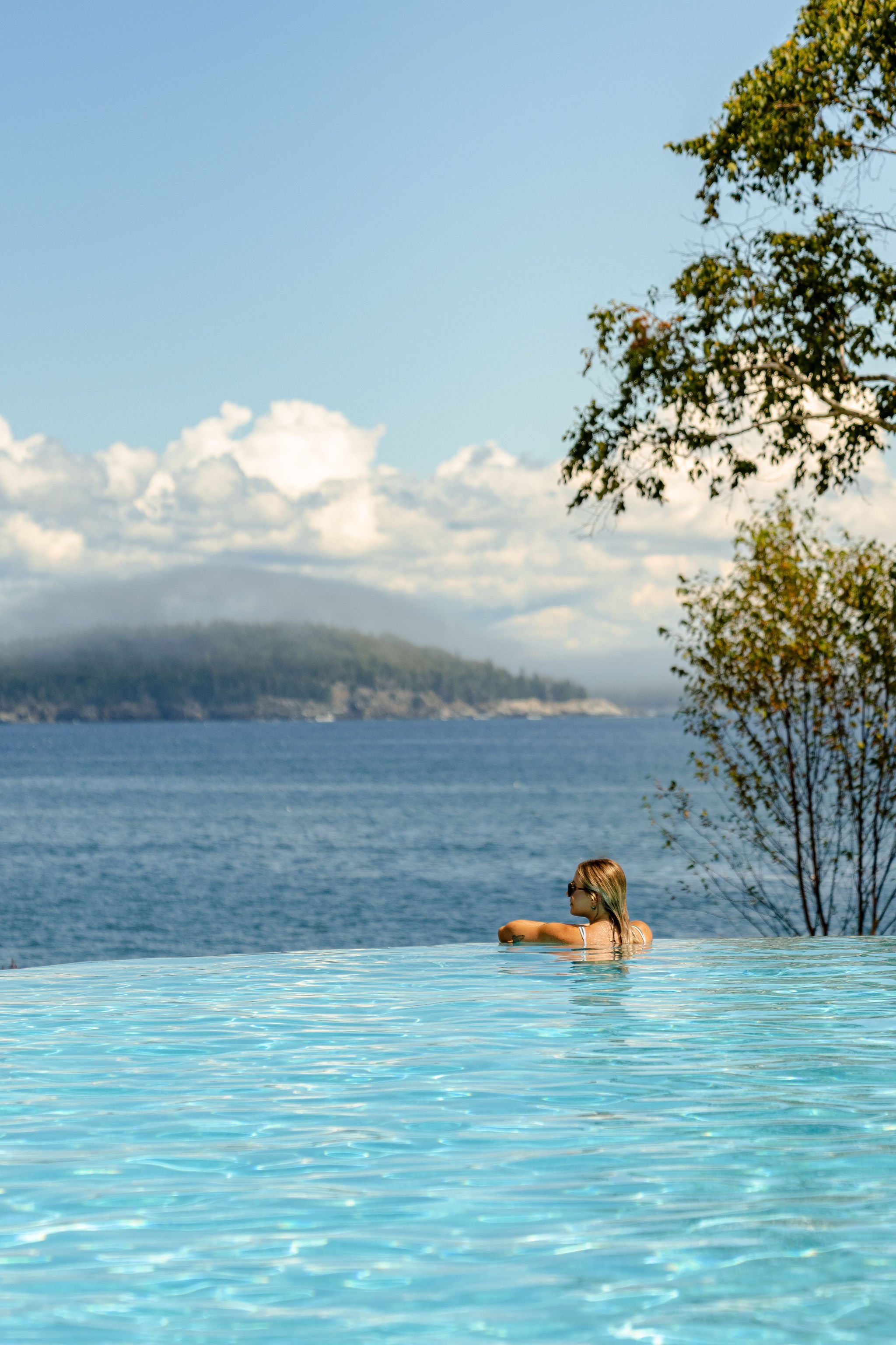 A woman relaxing in an infinity pool overlooking a lake or ocean, with trees and hills in the background under a partly cloudy sky.