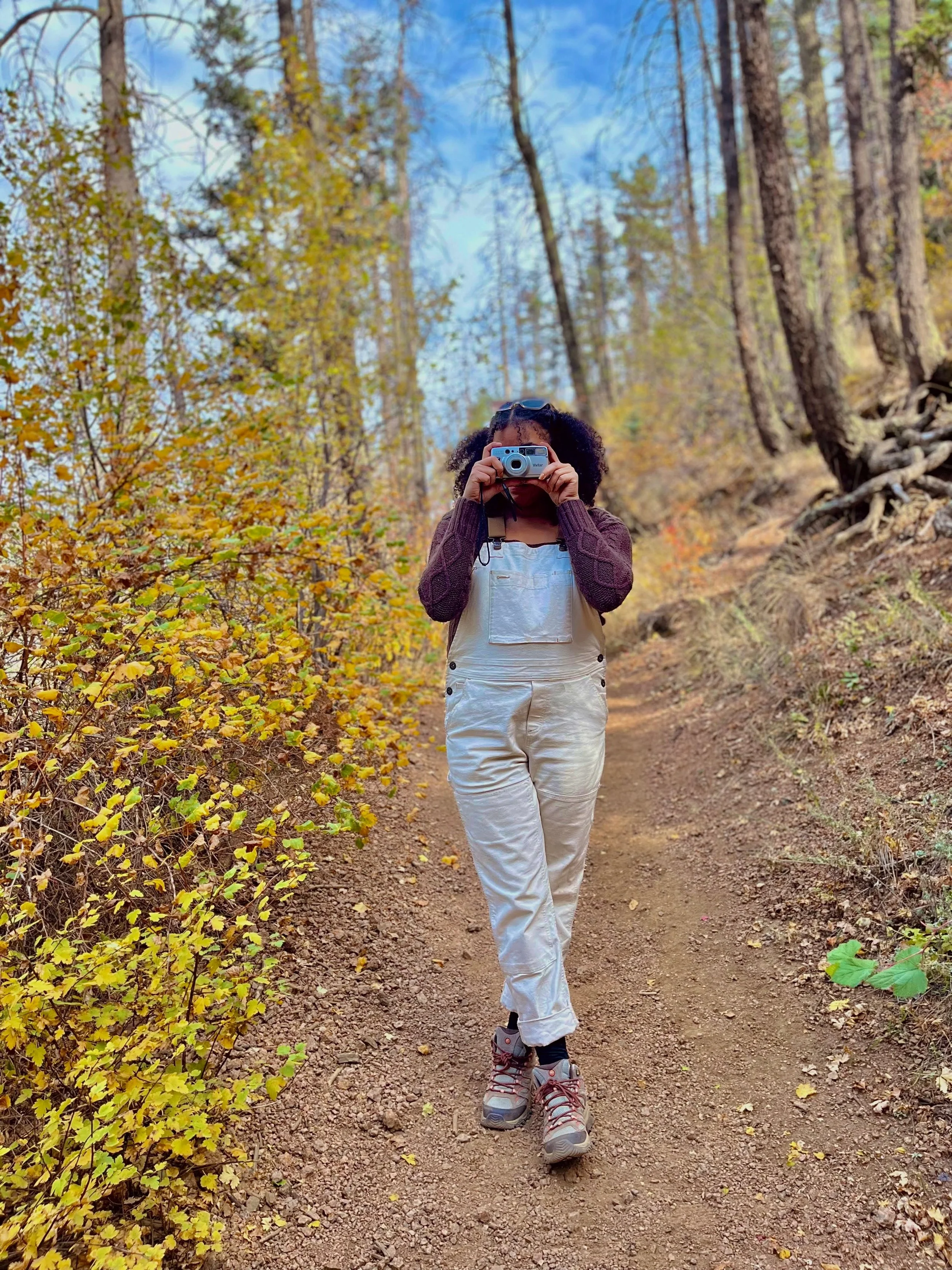 A woman on a hiking trail in a forest taking a photo with a camera, surrounded by autumn foliage and trees.