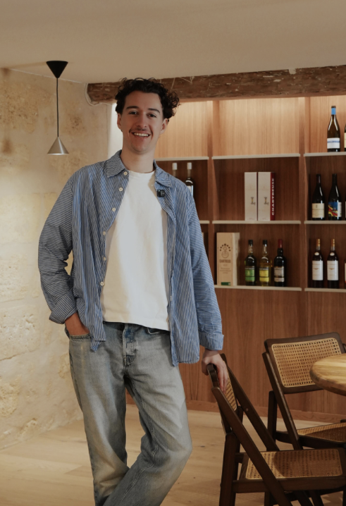 Un homme souriant se tenant dans une pièce avec des étagères en bois contenant des bouteilles de vin.