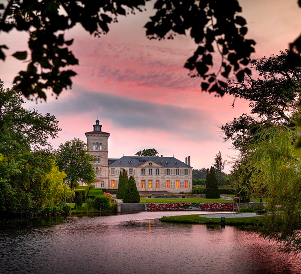 Un château historique entouré d'eau et de jardins, avec un ciel au coucher de soleil rose et violet.