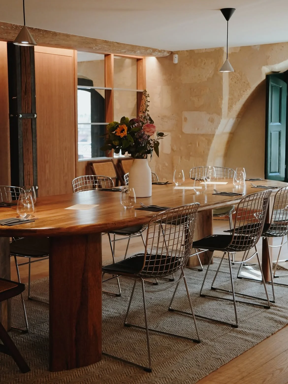 Salle à manger élégante avec une grande table en bois, des chaises en métal, un vase de fleurs, et un décor intérieur avec des murs en pierre et une fenêtre verte.