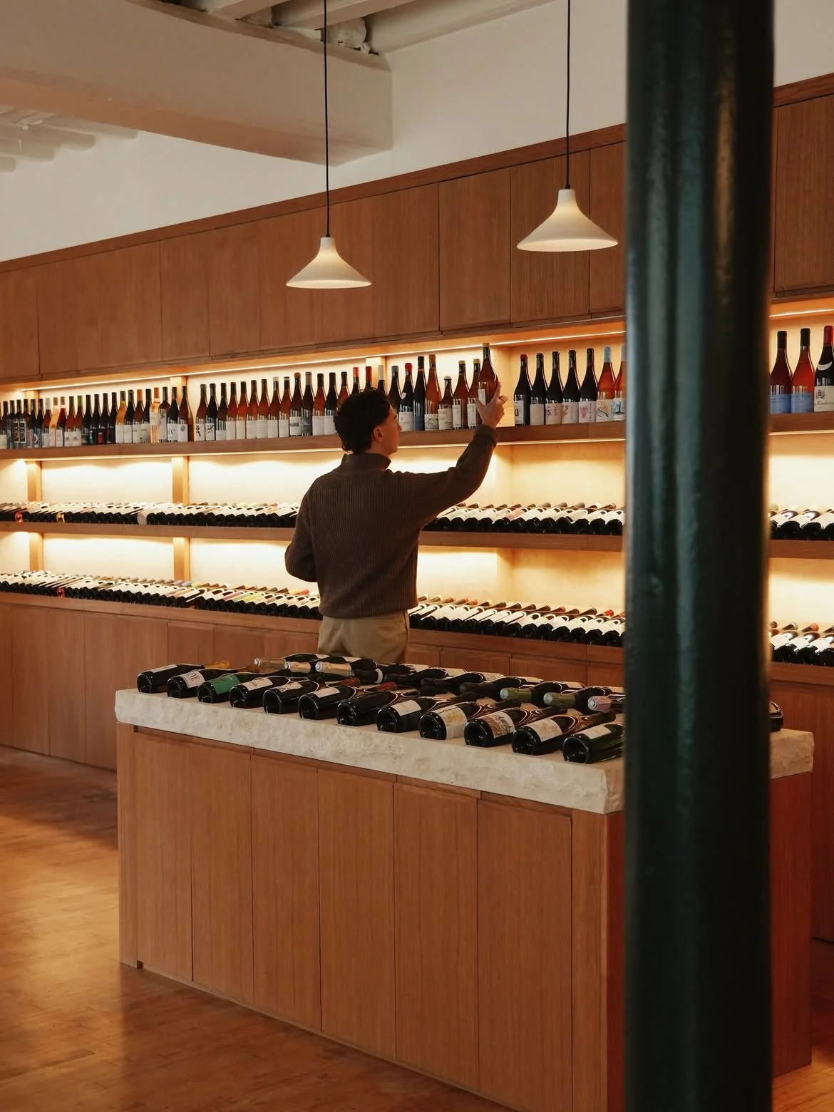 Une personne dans une boutique de vins, en train de sélectionner une bouteille sur une étagère en bois, entourée de nombreuses bouteilles de vin alignées.