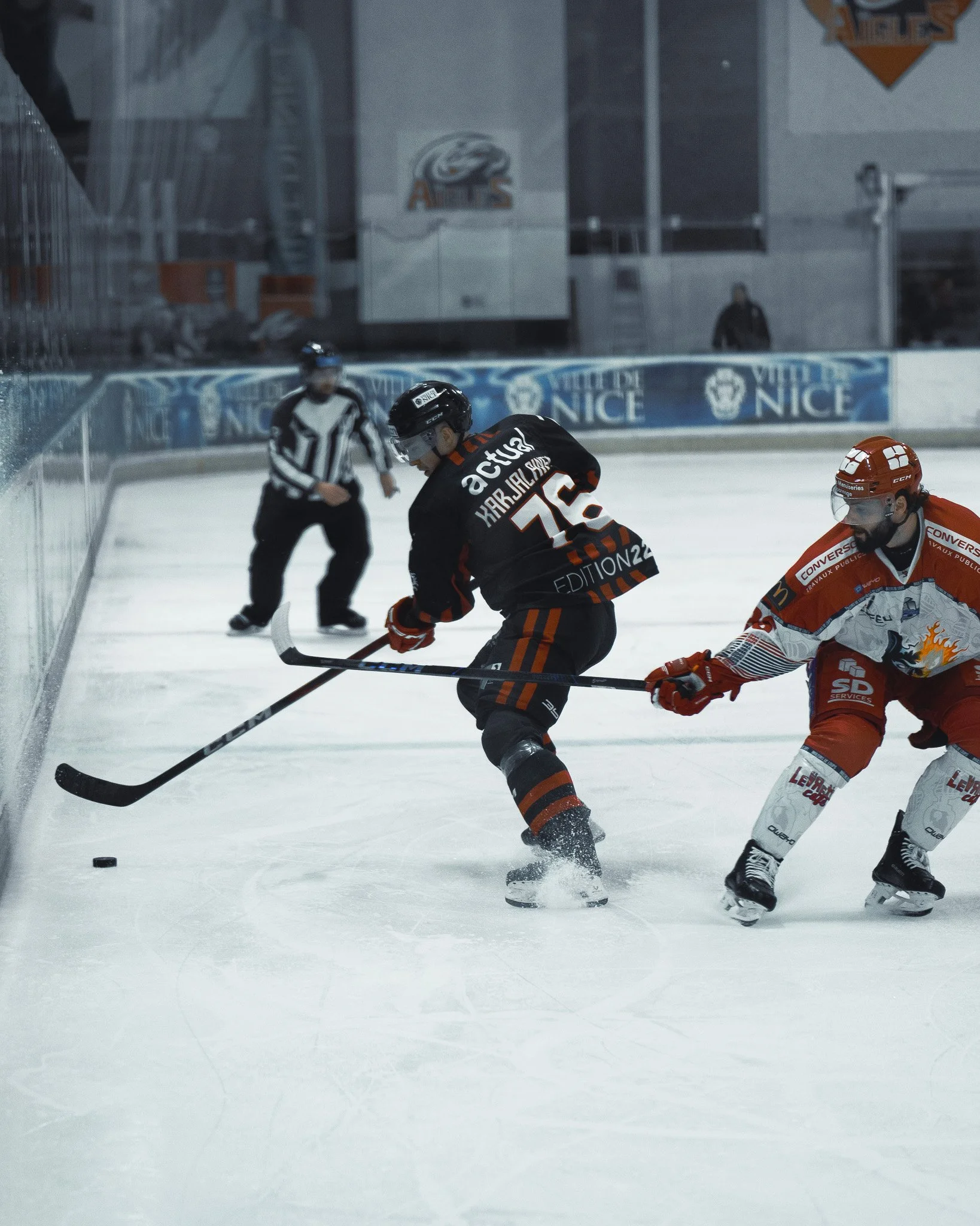 Deux joueurs de hockey sur glace en action lors d'un match, avec un arbitre observant en arrière-plan.