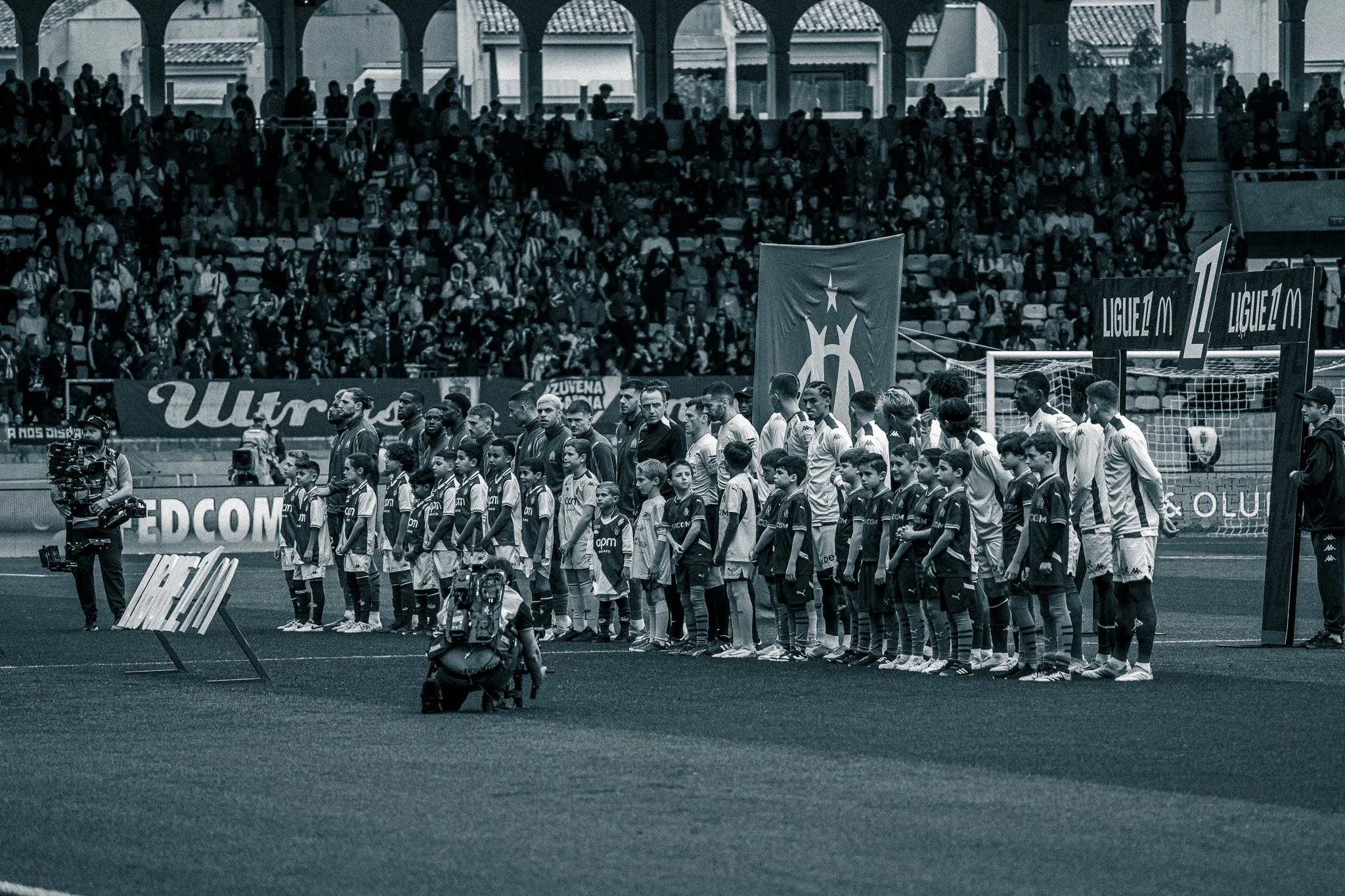 Une ligne de joueurs et d'enfants, probablement lors de l'avant-match, dans un stade de football avec un public en arrière-plan.