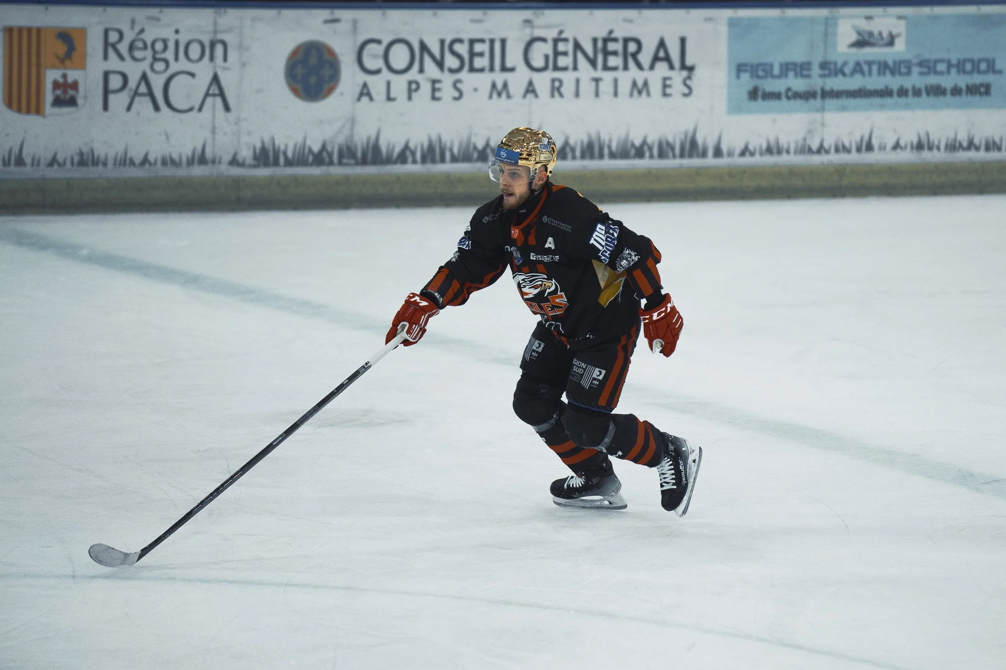 Joueur de hockey sur glace sur une patinoire, portant un casque doré et un équipement noir, s'apprêtant à frapper le palet.
