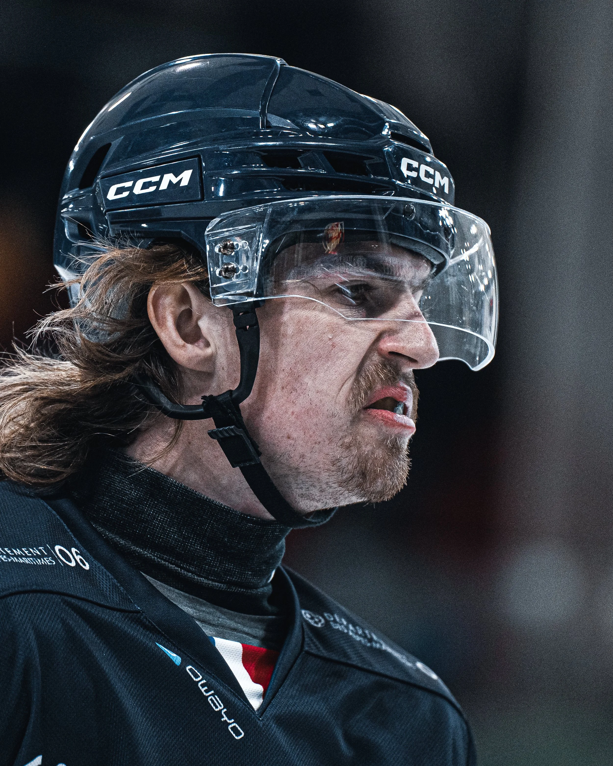 Un homme portant un casque de hockey avec une visière en plastique, portant un uniforme noir avec des logos et des numéros, regarde avec sérieux ou concentration.