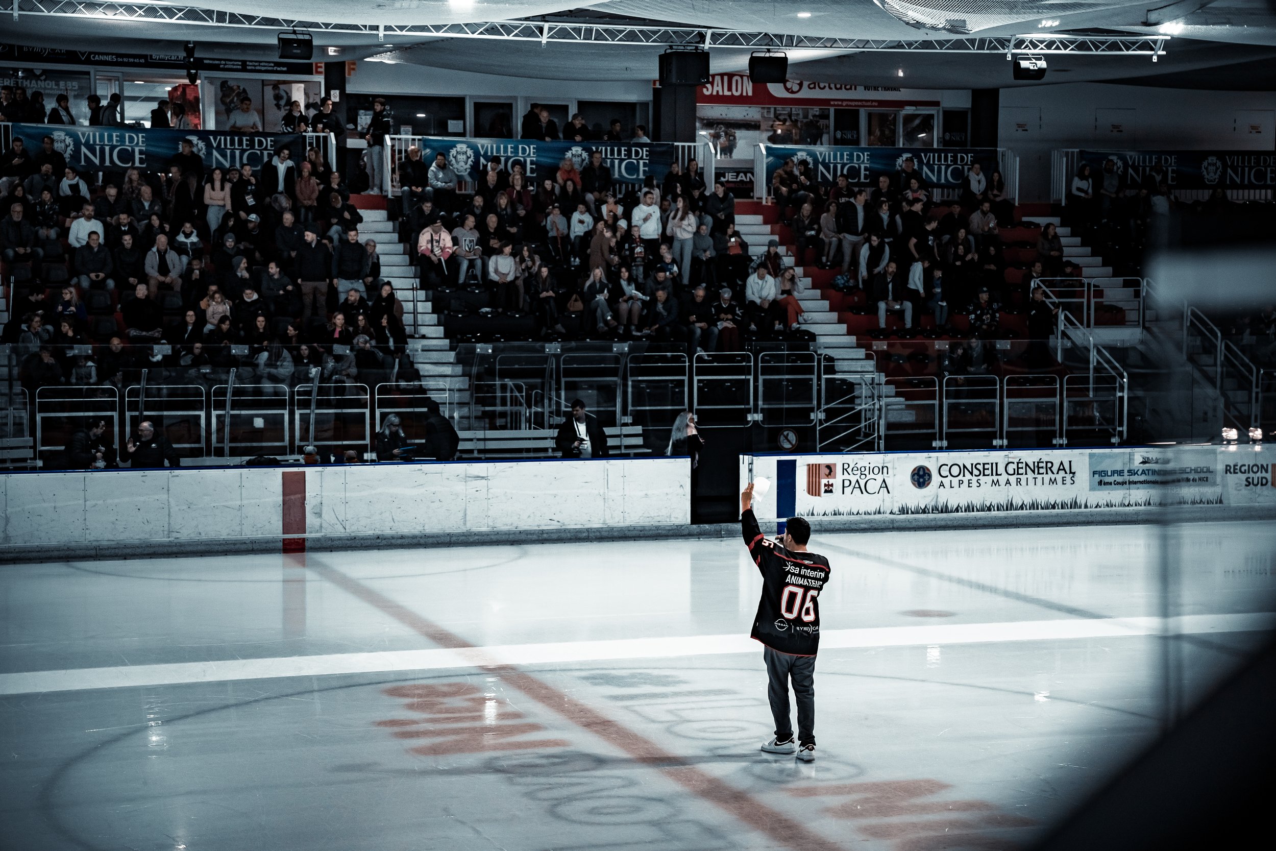 Personne sur une patinoire saluant une foule dans une arène de hockey, avec des spectateurs assis dans les gradins et des bannières affichant "Ville de Nice" en arrière-plan.