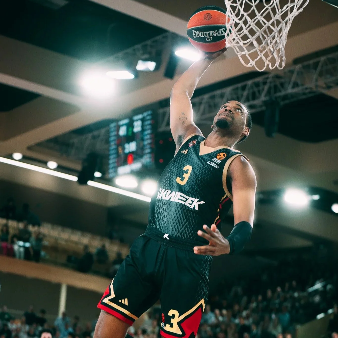 Un joueur de basketball en tenue noire avec le numéro 3, sautant pour la balle dans le panier, dans une salle de sport lumineuse avec spectateurs en arrière-plan.
