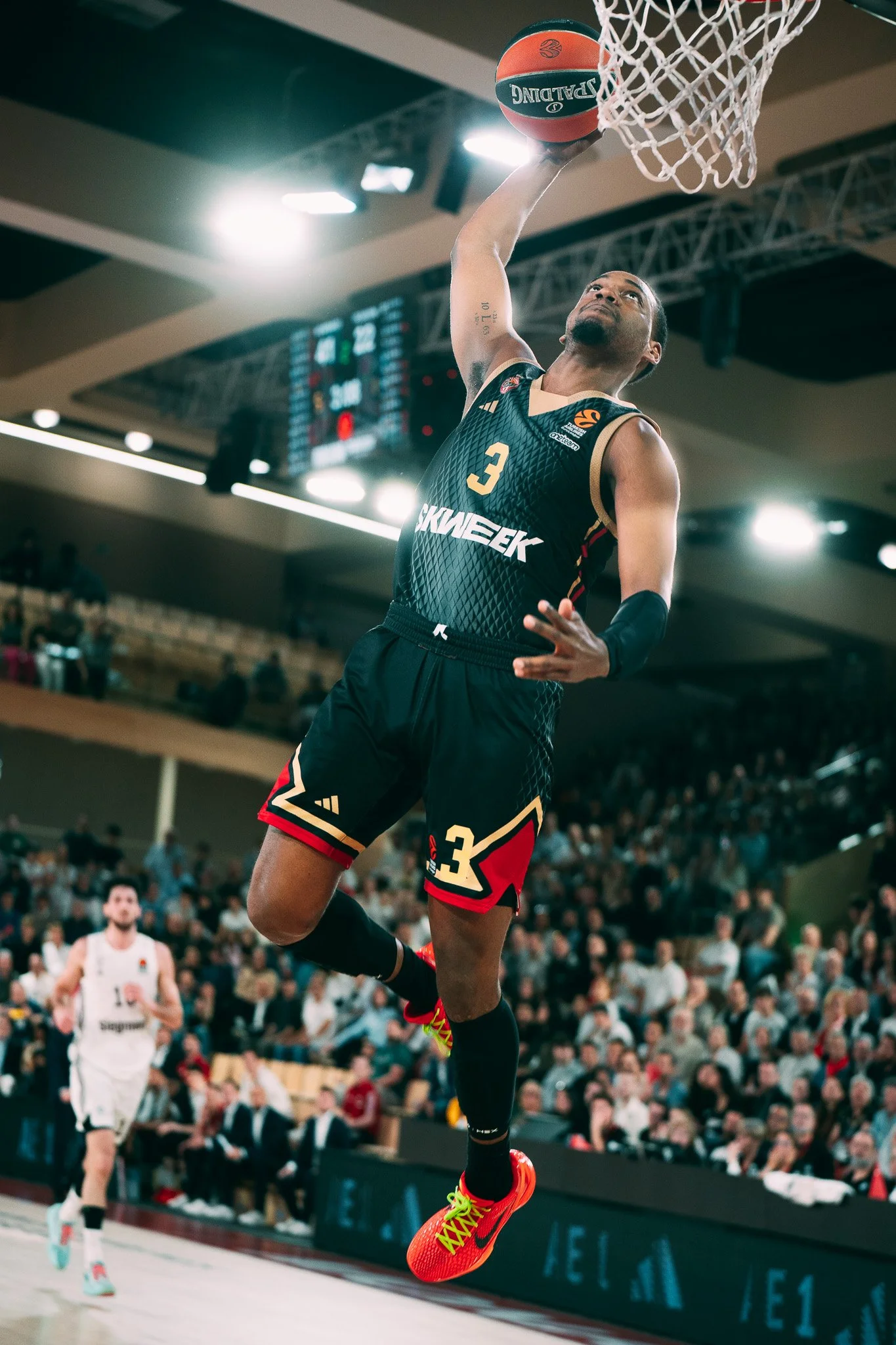 Un joueur de basket en pleine action lors d'un match, sautant vers le panier pour dunker, portant un uniforme noir avec le numéro 3, dans une salle bondée. Un ballon de basketball est en suspension près du panier.