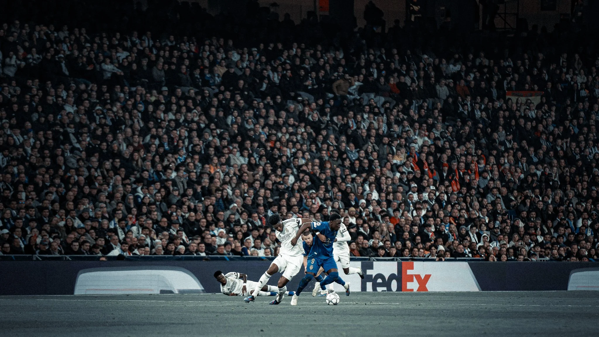 Match de football avec plusieurs joueurs en action sur le terrain, un grand public dans les gradins en arrière-plan, publicité de FedEx visible sur la bande de bordure du terrain.
