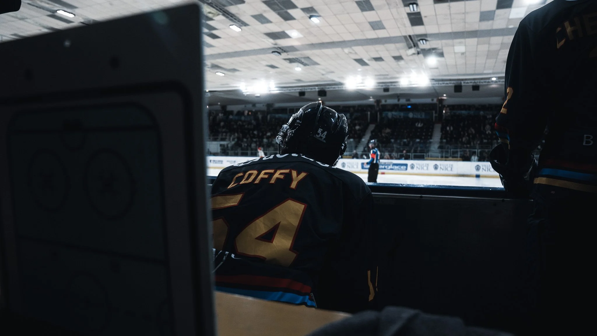 Joueur de hockey regardant le match depuis la zone de la patinoire, portant un casque et un maillot avec le nom 'COFFY' et le numéro 14.