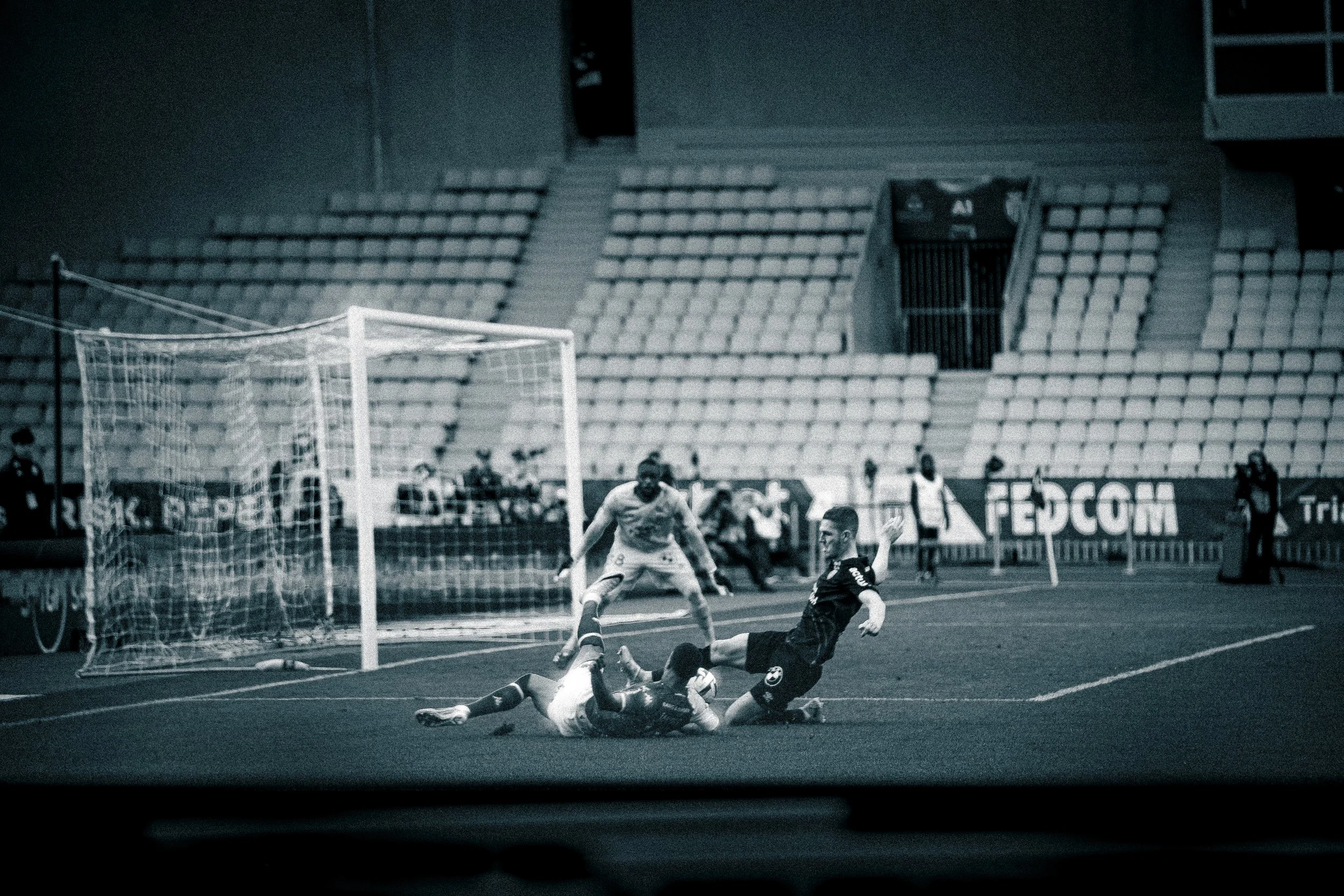 Une scène de match de football vue à travers une vitre, montrant un joueur en noir tentant de marquer un but tandis que le gardien et un défenseur tentent de l'arrêter, dans un stade presque vide.