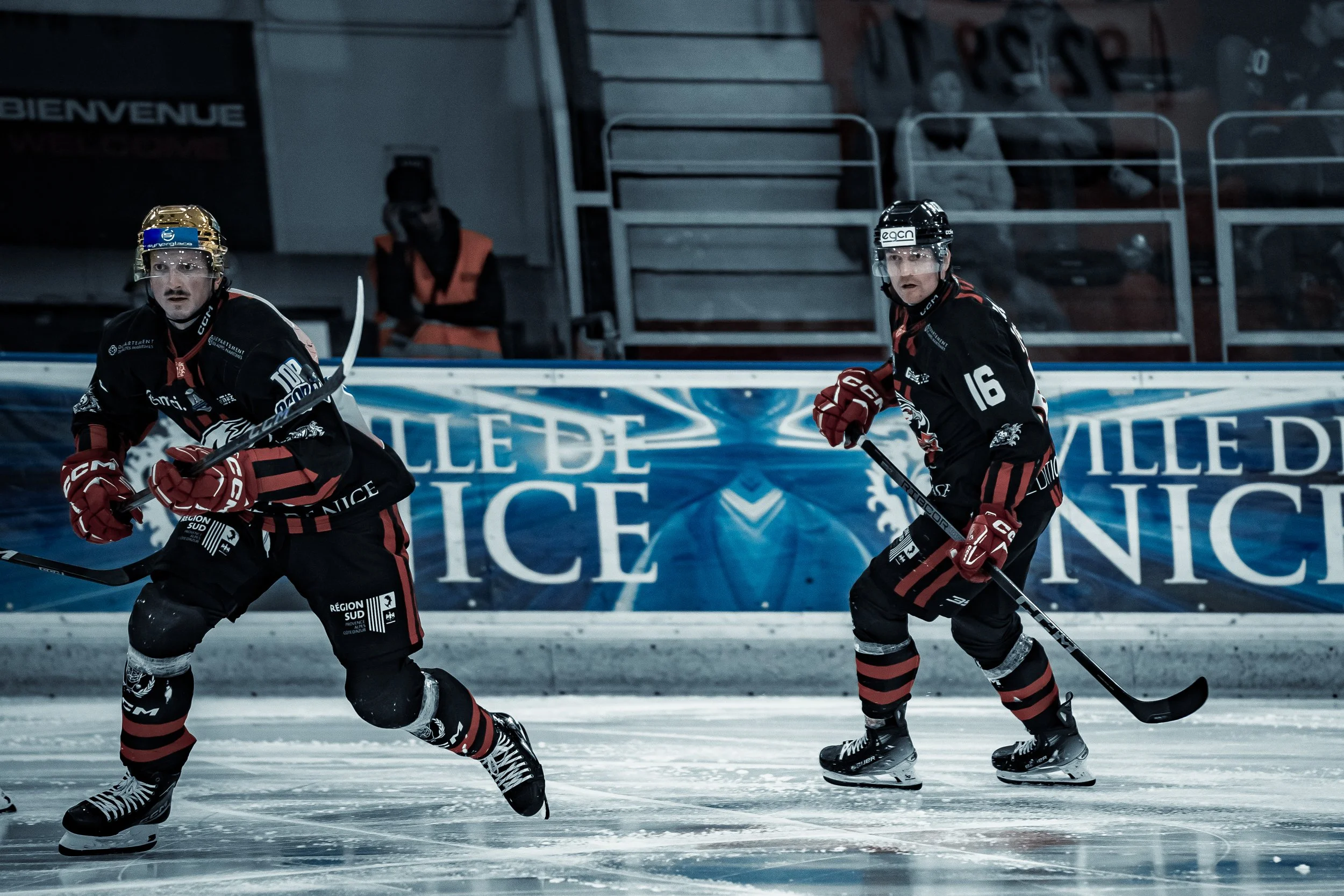 Deux joueurs de hockey sur glace sur une patinoire, portant des uniformes noirs avec des accents rouges. Ils tiennent des bâtons de hockey et patinent pendant un match. Une bannière en arrière-plan mentionne "VILLE DE NICE".