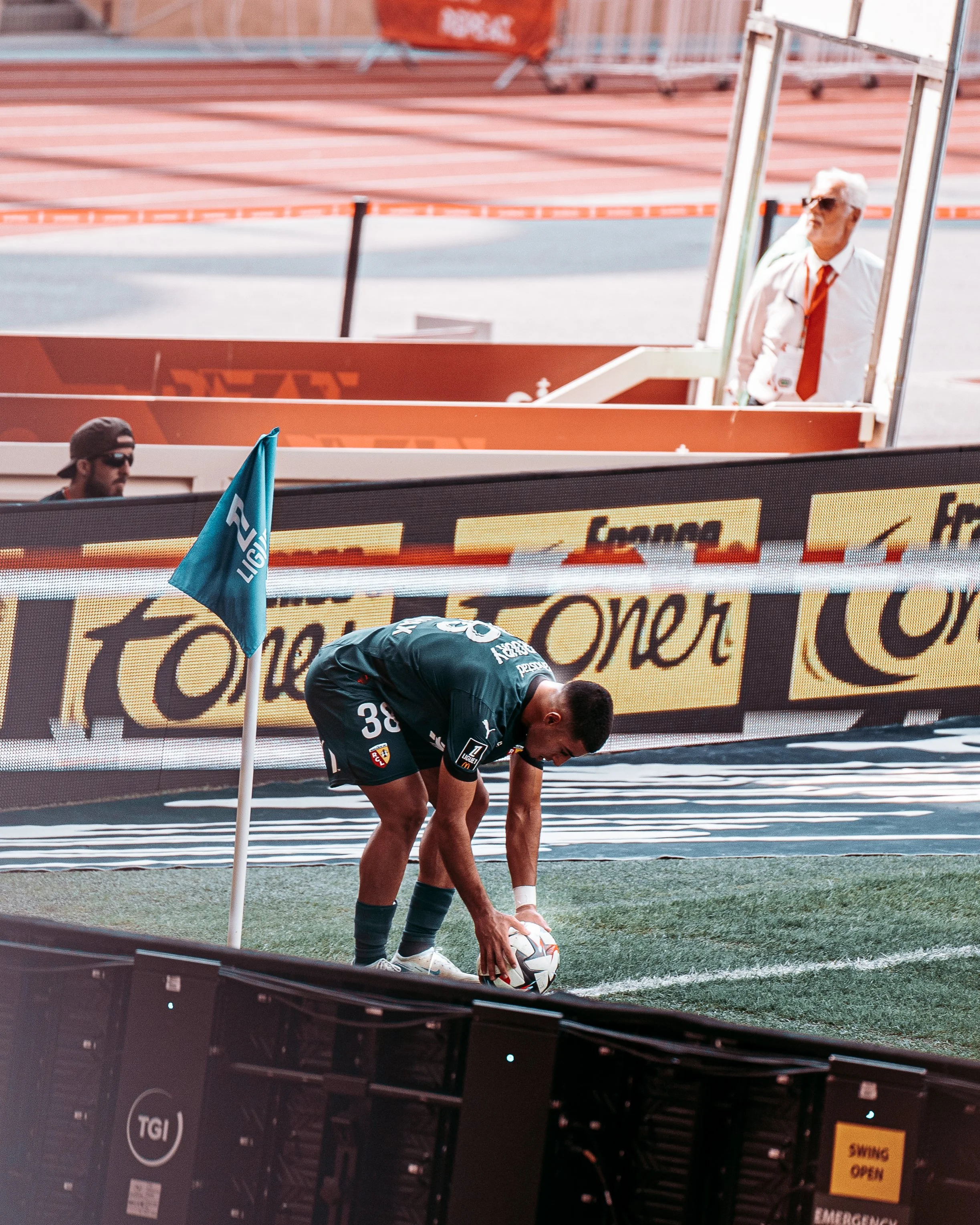 Un footballeur en maillot vert s'apprête à tirer un corner sur un terrain de football, avec un drapeau de coin de la Ligue 1 et des spectateurs en arrière-plan.