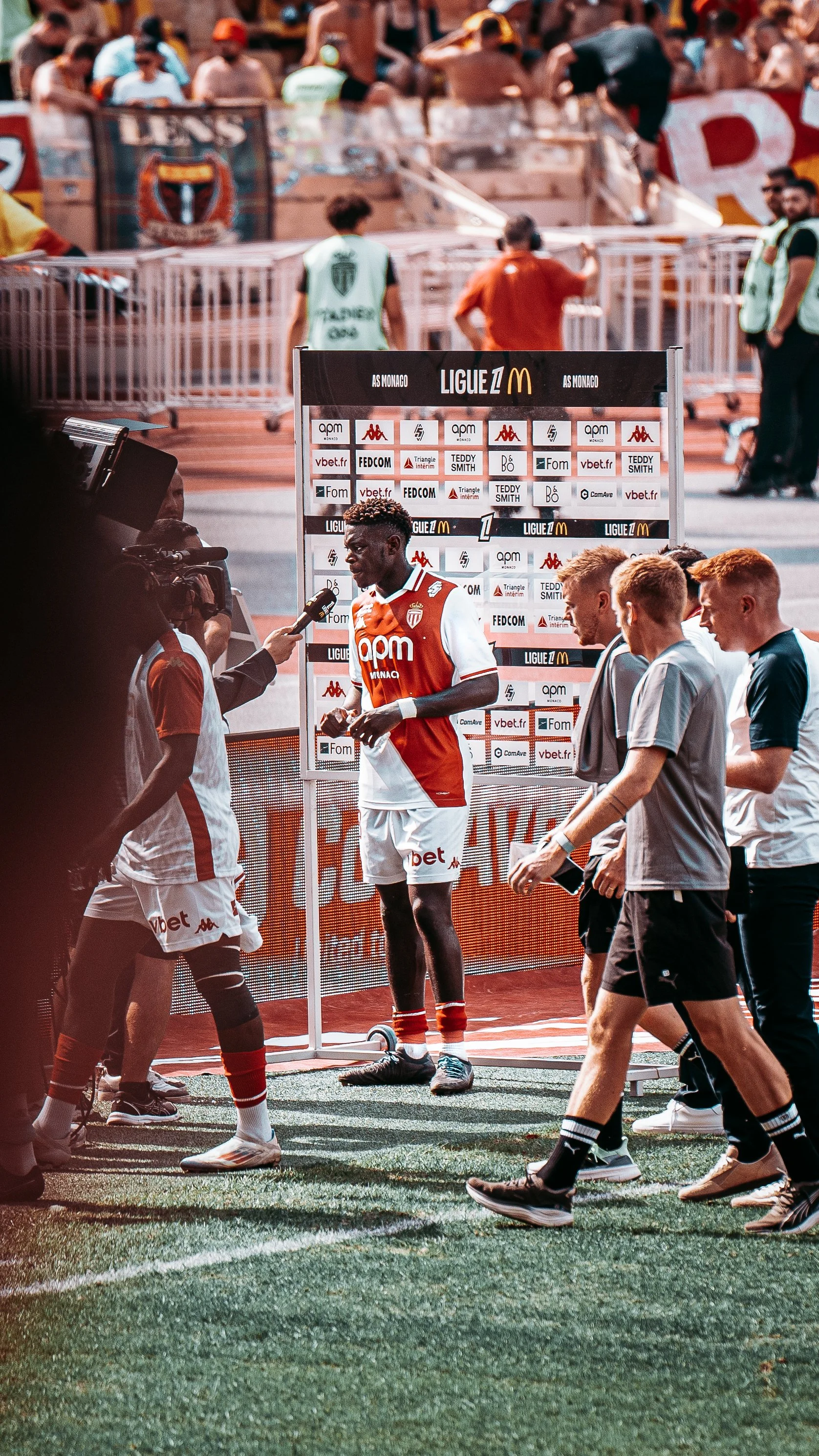 Interviewer un joueur de football sur le terrain après un match, avec un tableau de sponsors et plusieurs personnes en arrière-plan.