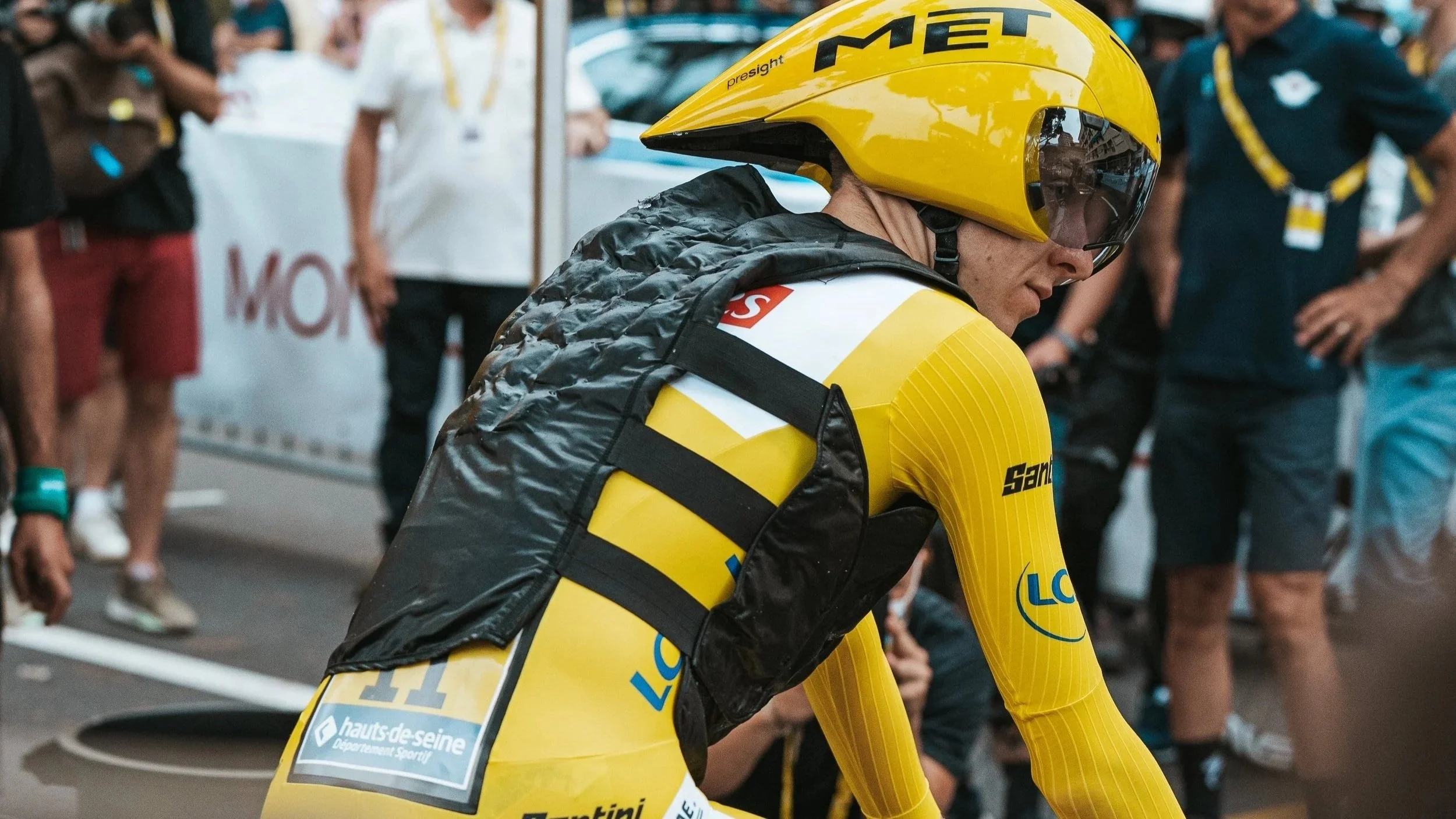 Un cycliste en tenue jaune et casque jaune, prêt à courir, au départ d'une course, avec des spectateurs en arrière-plan