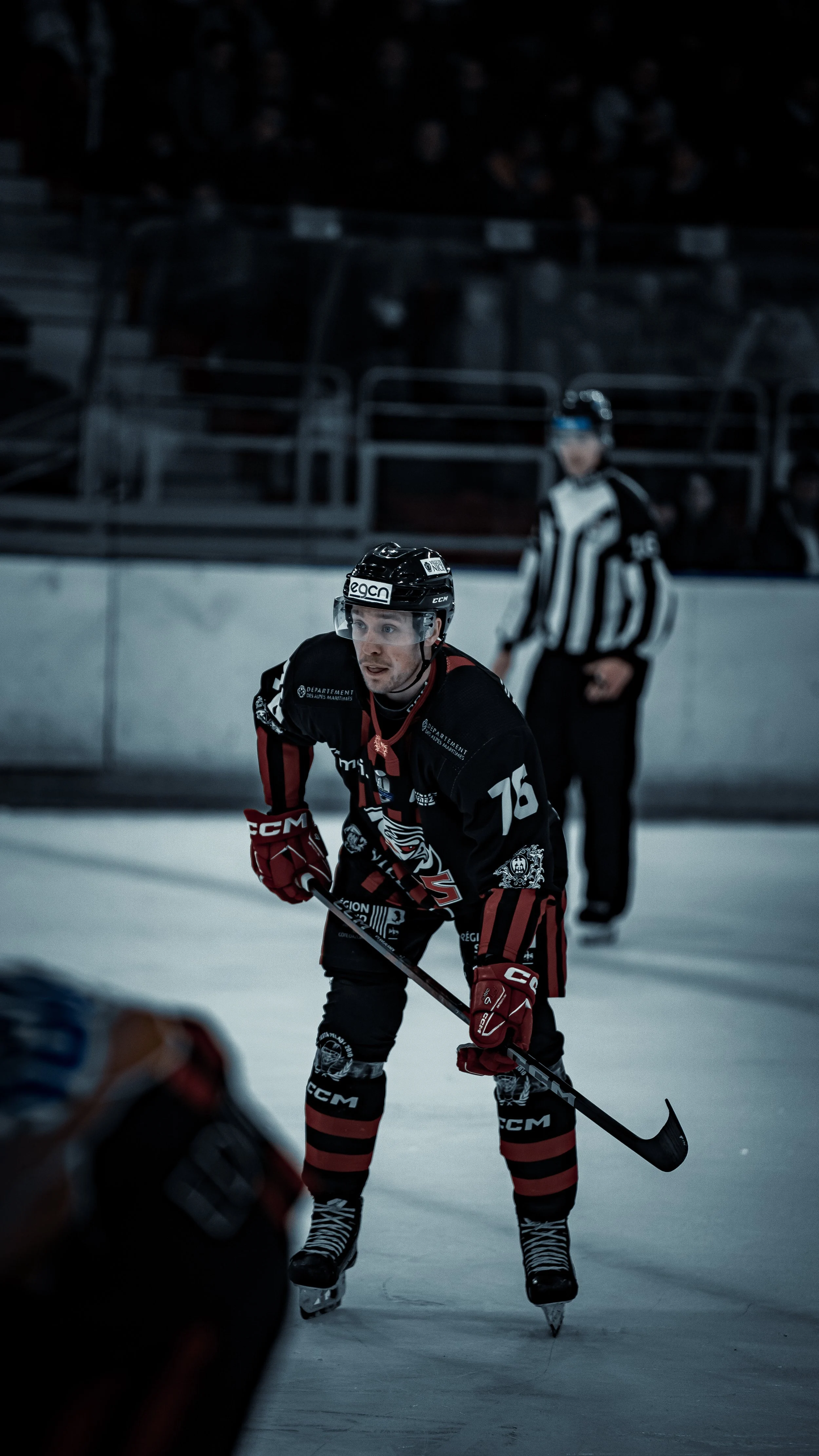 Joueur de hockey sur glace en action sur la patinoire, portant un maillot noir et rouge avec le numéro 76. Un arbitre flou en arrière-plan.