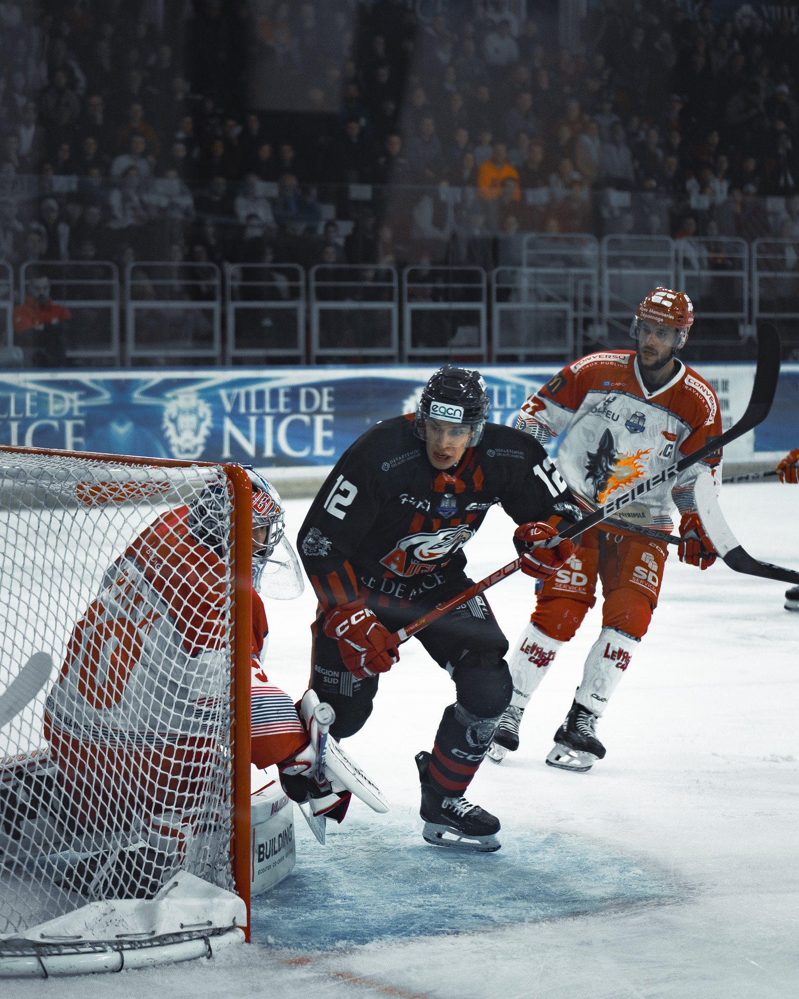 Joueurs de hockey sur glace en action devant le but lors d'un match avec spectateurs en arrière-plan.