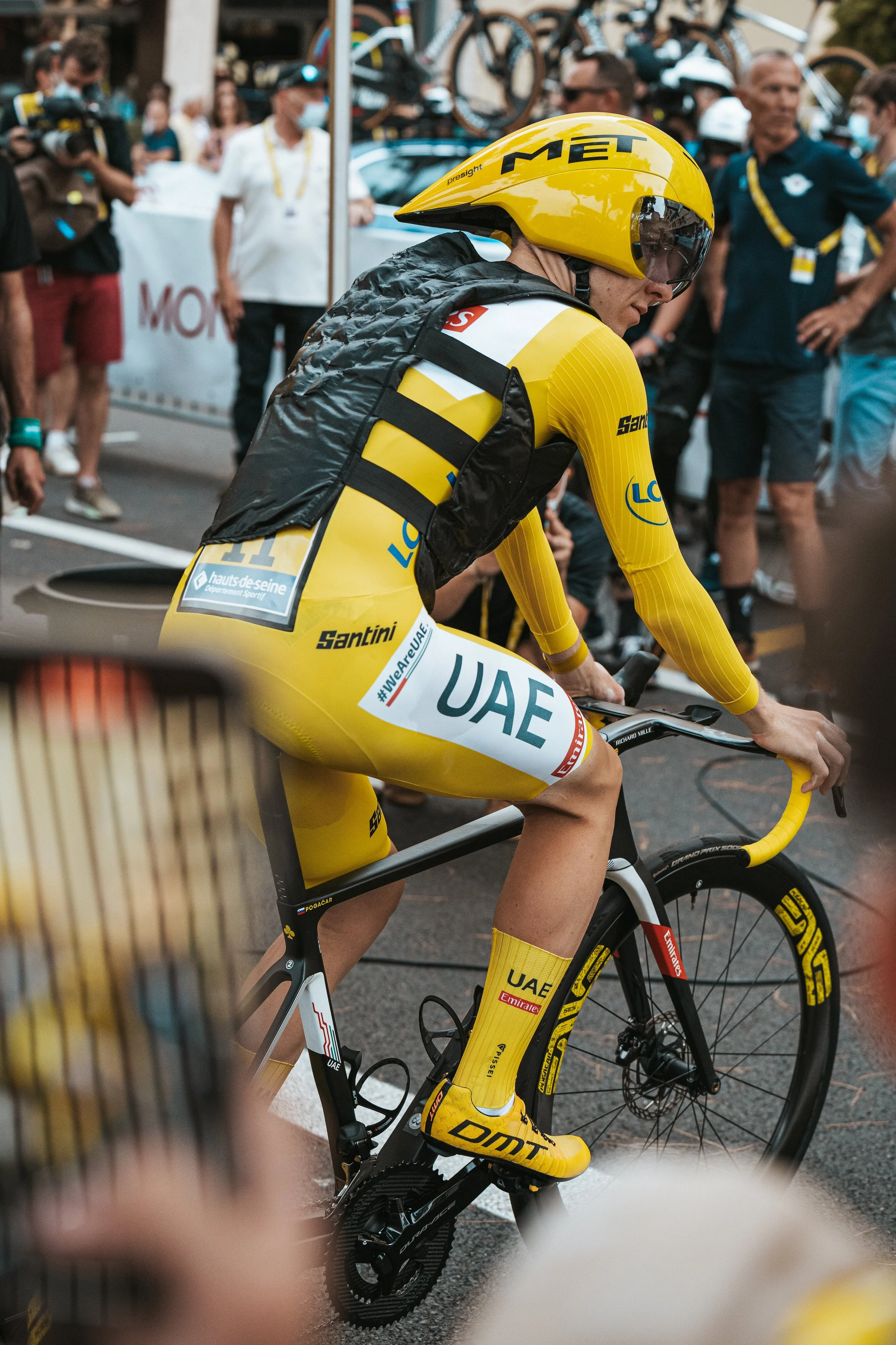 Cycliste portant un maillot et des chaussures jaunes, assis sur un vélo lors d'une course ou d'un événement sportif avec des spectateurs en arrière-plan.