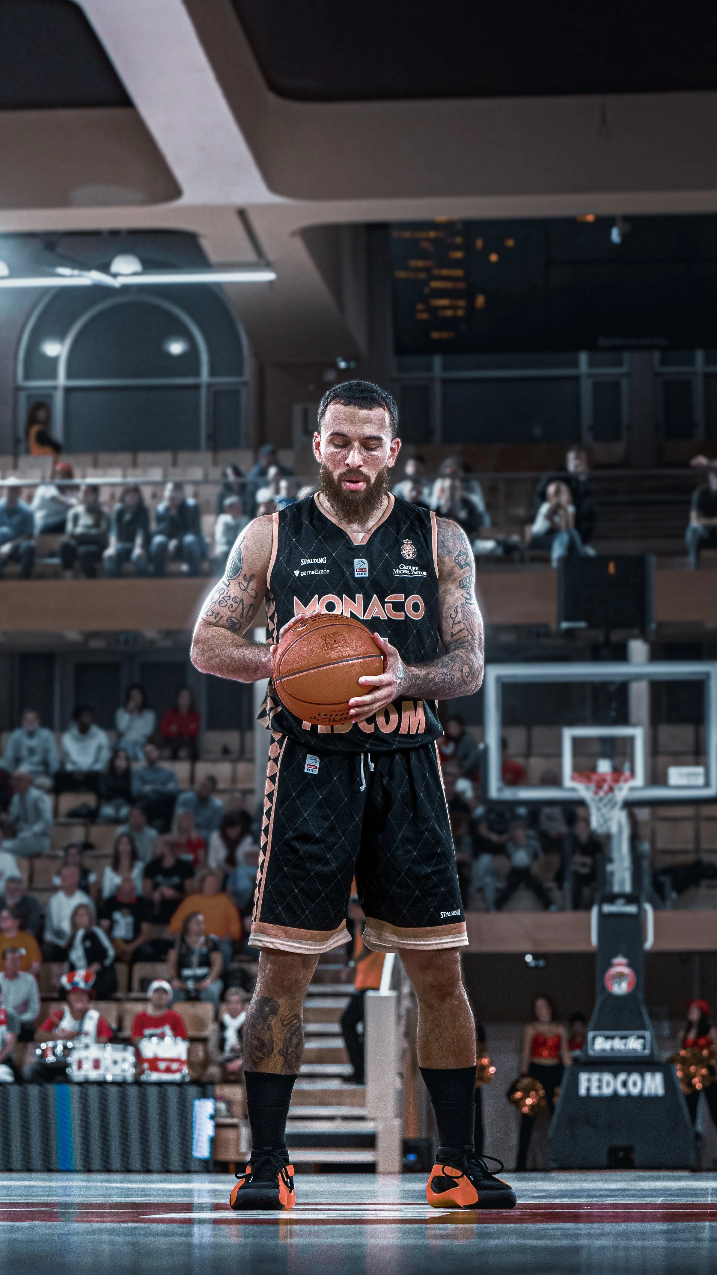 Un joueur de basket-ball en tenue noire avec le mot 'MONACO' écrit dessus, tenant un ballon de basket, sur un terrain de basket dans une salle bondée.