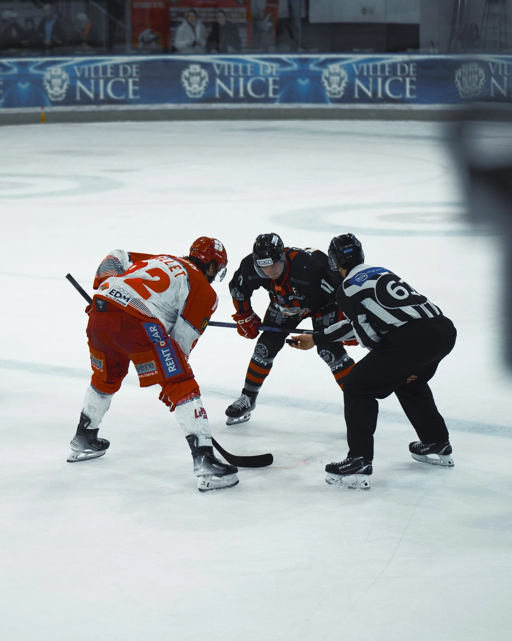 Deux joueurs de hockey sur glace s'affrontent lors d'une remise en jeu, avec un arbitre entre eux, sur une patinoire avec l'inscription "Ville de Nice" en arrière-plan.