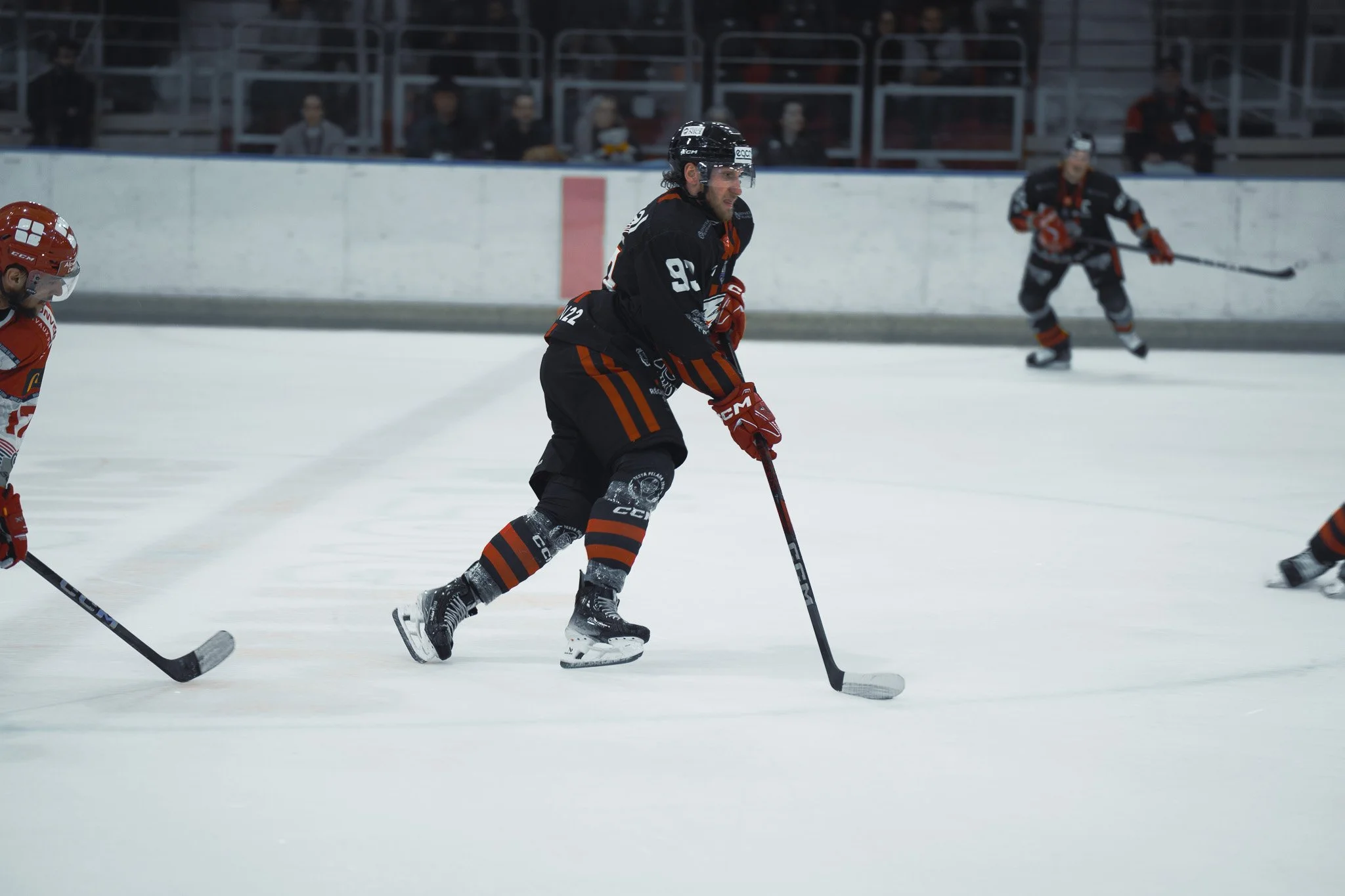 Joueurs de hockey sur glace en action sur la patinoire, portant des uniformes noirs et rouges, avec des spectateurs en arrière-plan.