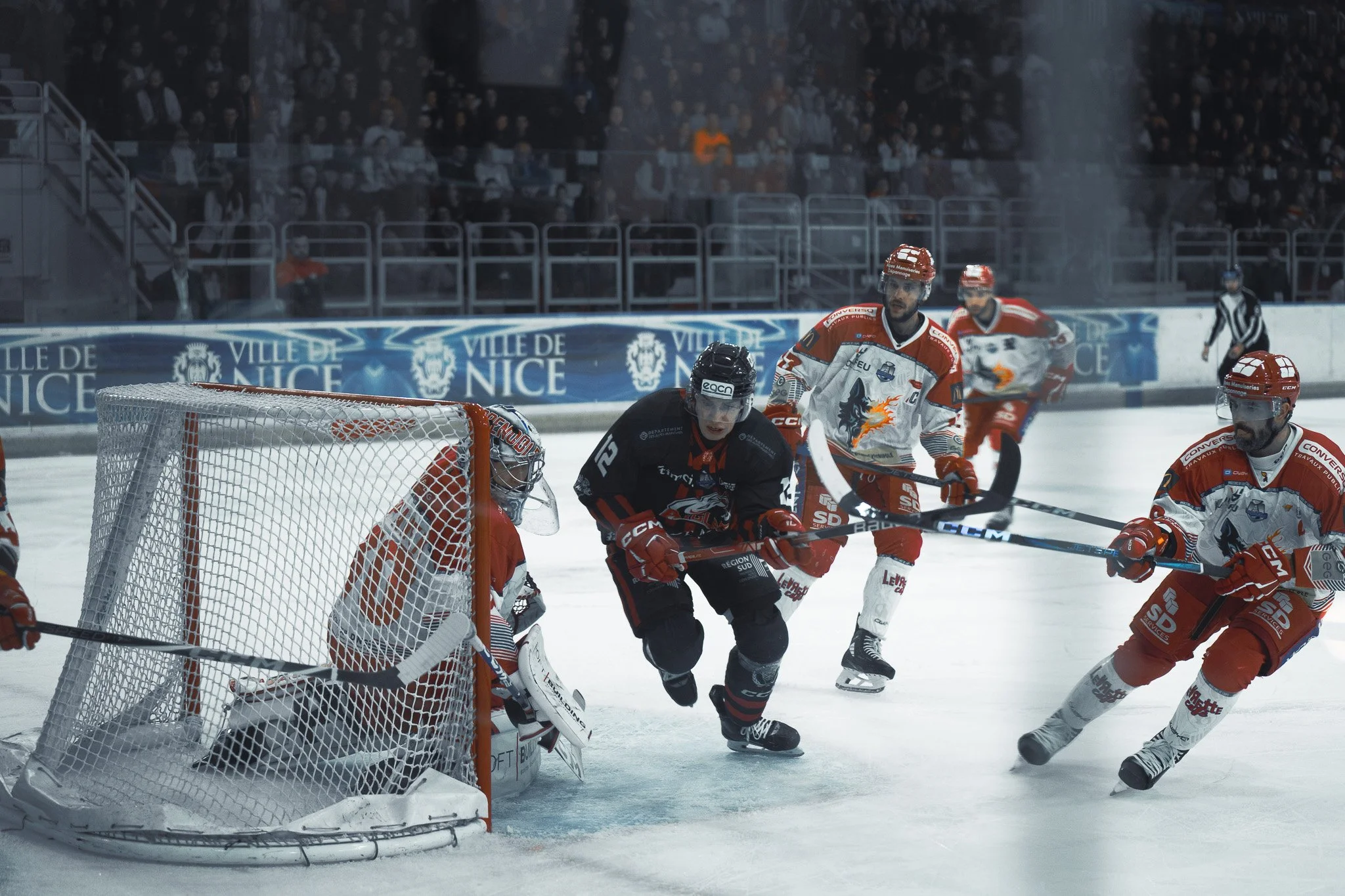 Match de hockey sur glace en cours, avec des joueurs en tenue rouge et noire sur la patinoire, à l'arrière-plan un panneau indiquant "Ville de Nice".
