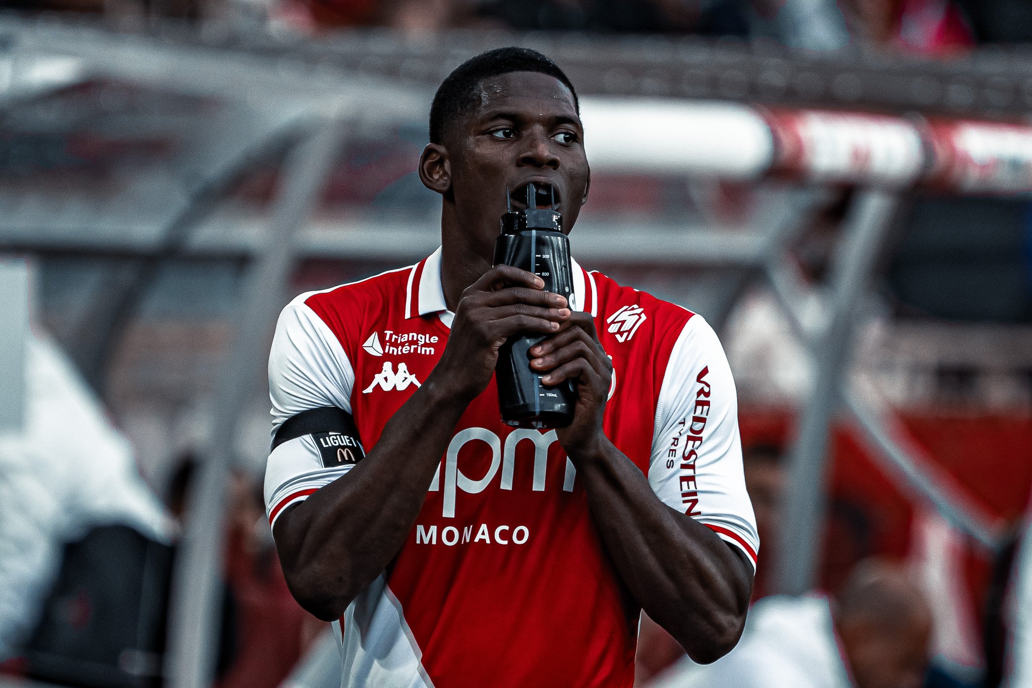 Un joueur de football en maillot rouge et blanc, portant un brassard noir, boit dans une bouteille d'eau sur le banc de touche d'un stade.