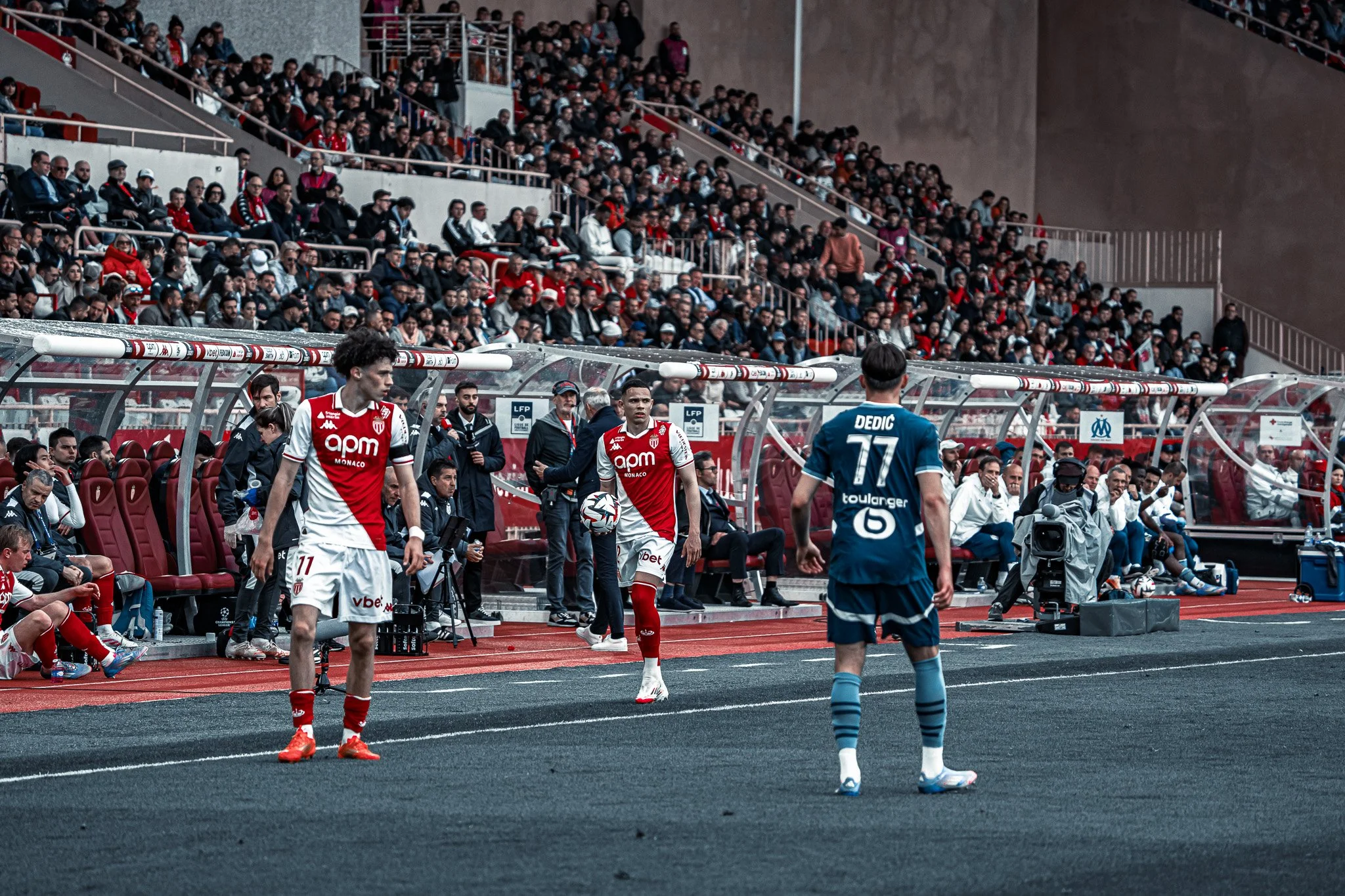 Match de football avec des joueurs en action, un public nombreux dans le stade, et des coéquipiers et entraîneurs sur le banc.