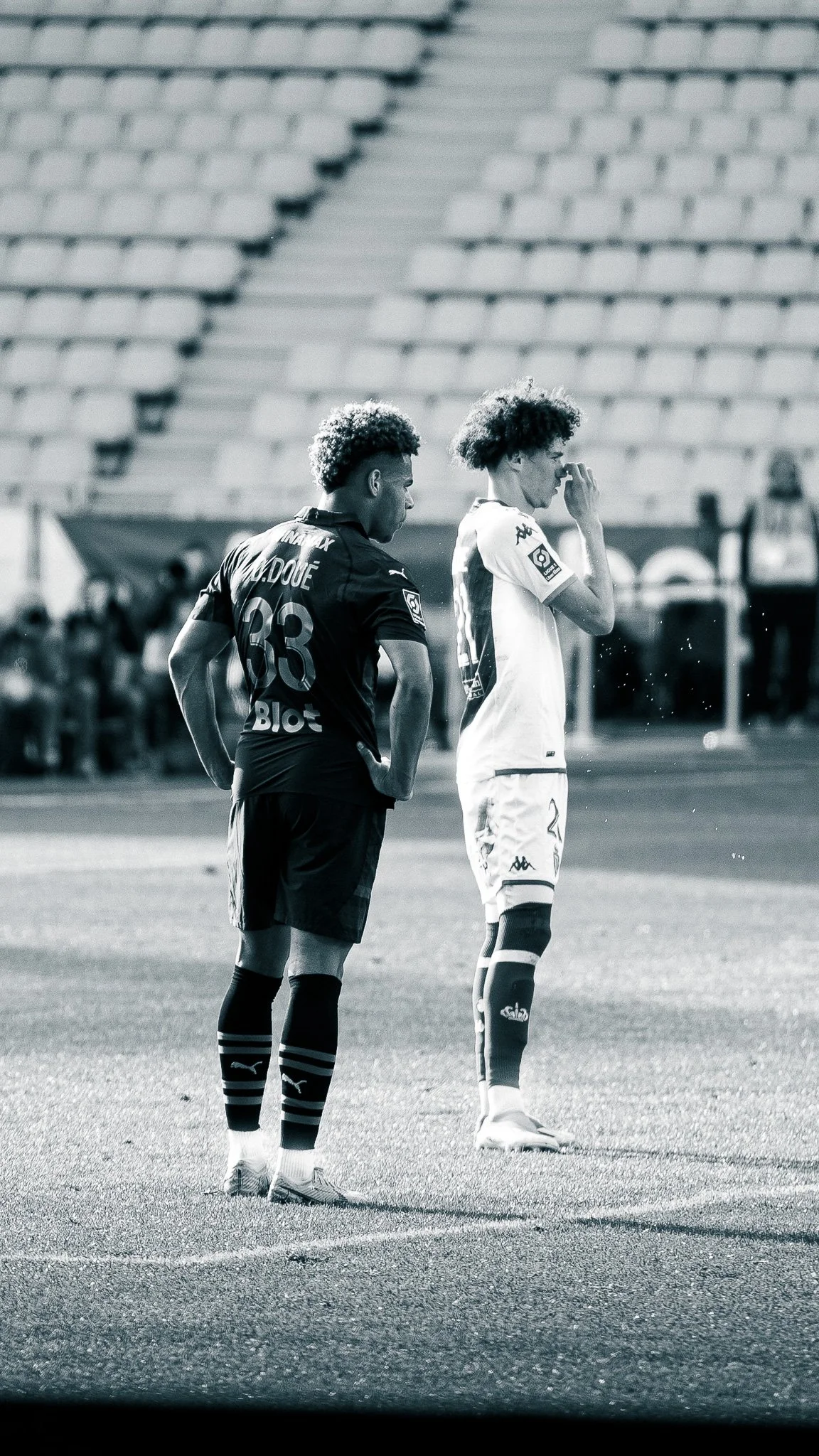 Deux joueurs de football, un en maillot noir et l'autre en maillot blanc, se tiennent sur un terrain de football vide, en pleine période avant-match ou pendant une pause.