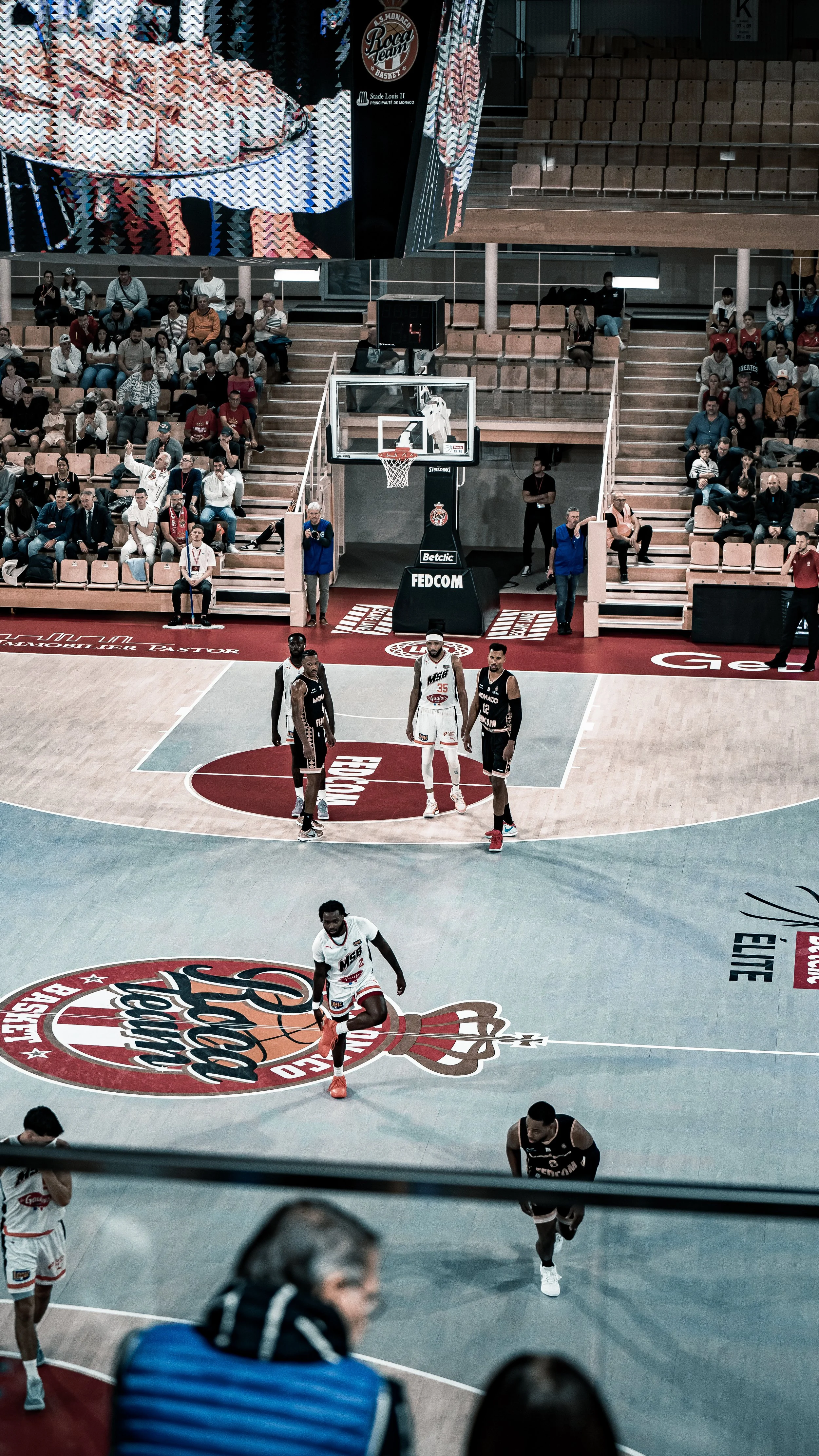 Match de basketball au stade Louis II avec joueurs en action et spectateurs dans les gradins, tableaux publicitaires et écran géant