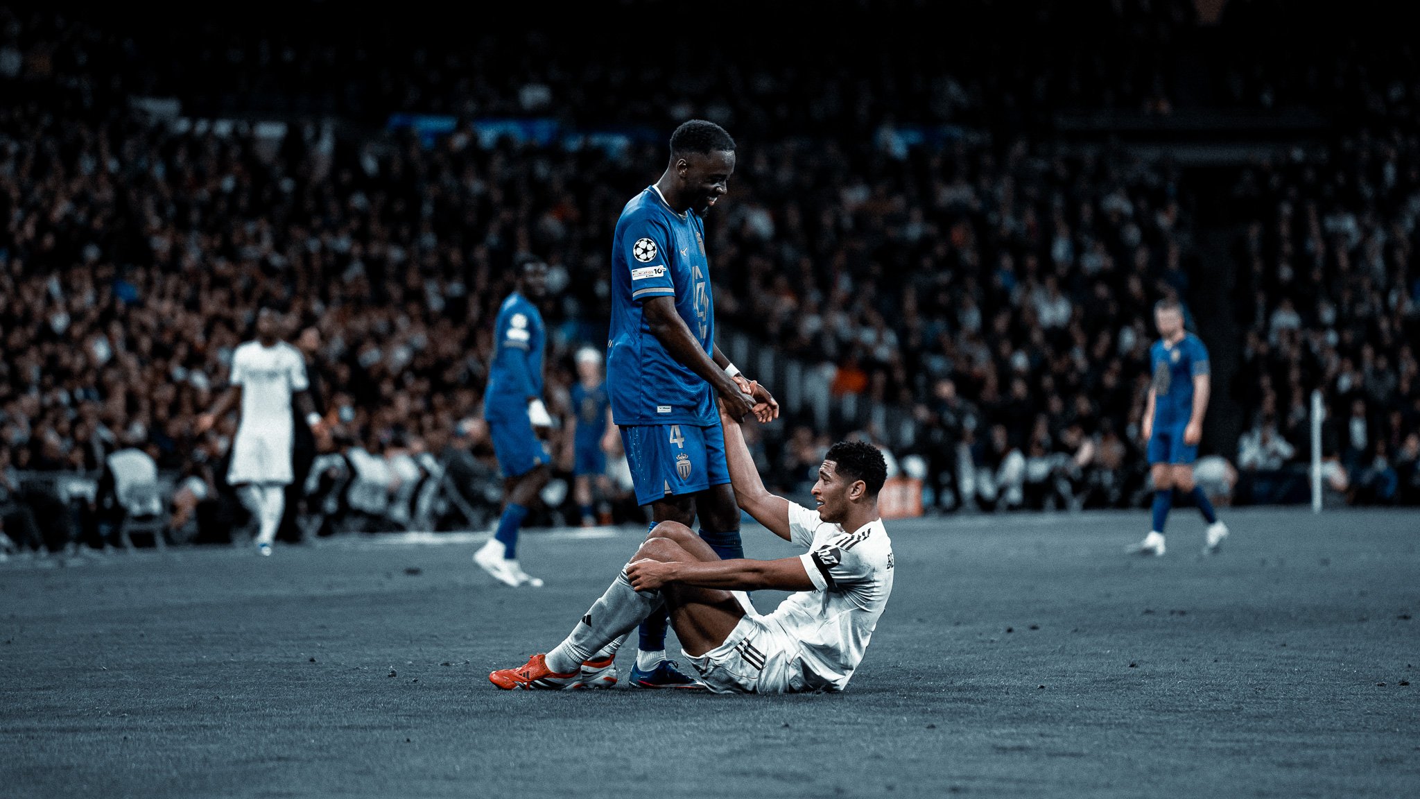 Deux joueurs de football, l'un en maillot blanc et l'autre en maillot bleu, en train d'interagir sur le terrain avec une foule nombreuse en arrière-plan.
