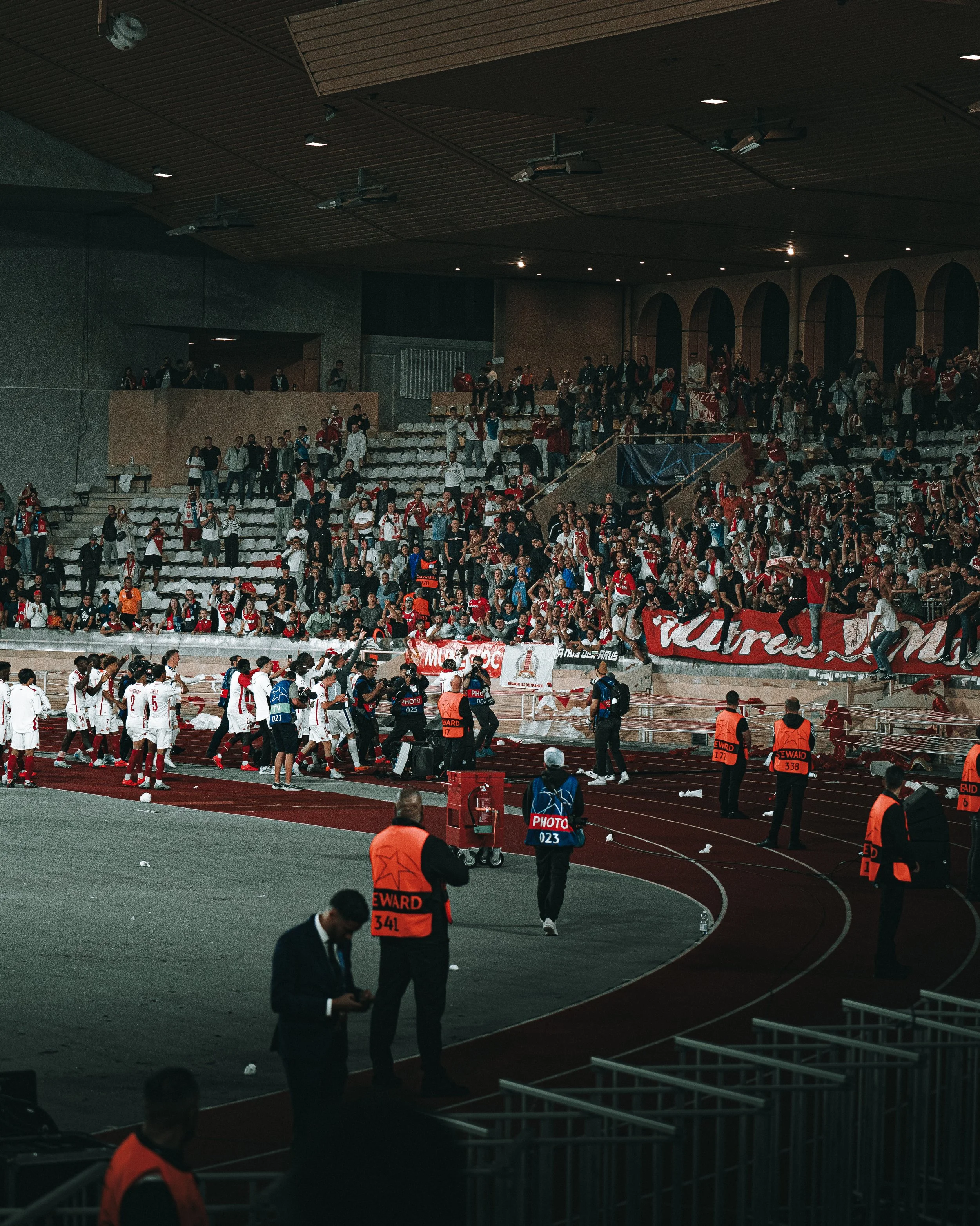 Spectateurs et joueurs dans un stade lors d’un match de football, certains joueurs en tenue blanche se dirigent vers le banc, security et photographes présents.