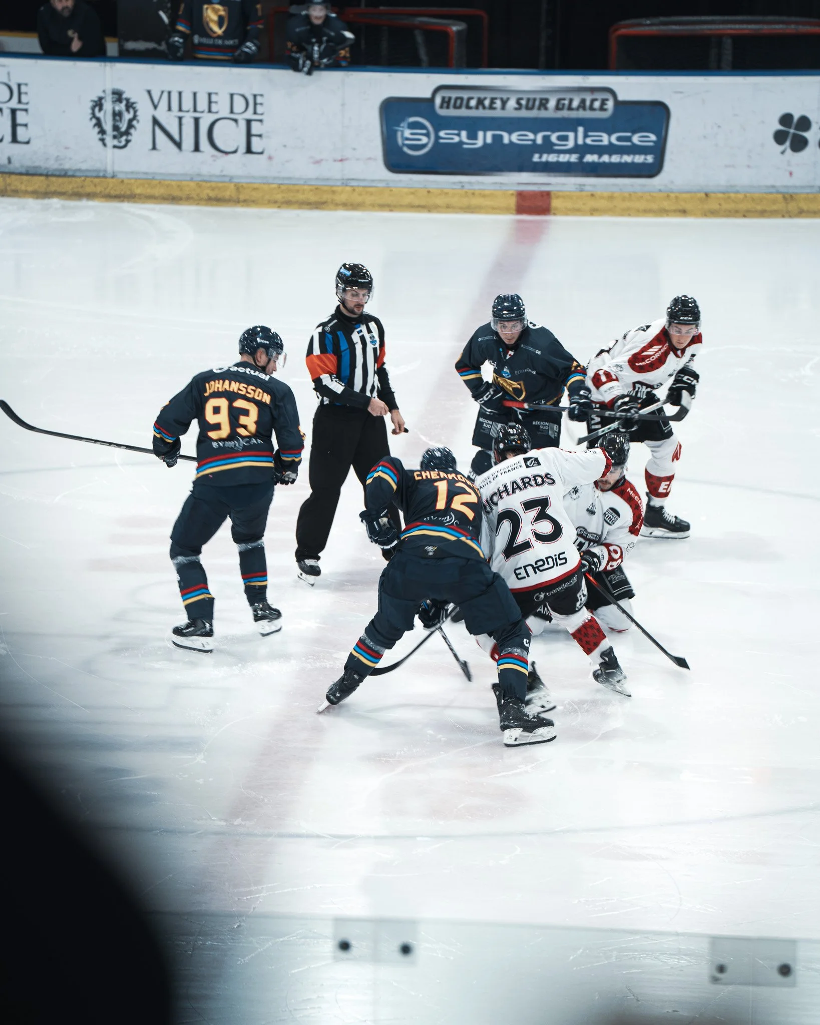 Match de hockey sur glace avec joueurs en action, un arbitre, et des panneaux publicitaires en arrière-plan.