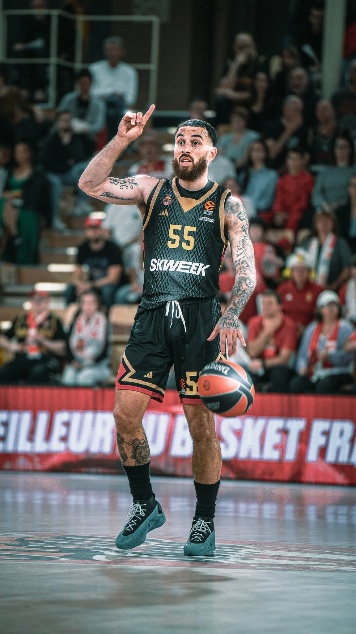 Joueur de basketball en train de faire une capture de balle lors d’un match, portant un maillot noir avec le numéro 55, avec un public en arrière-plan.