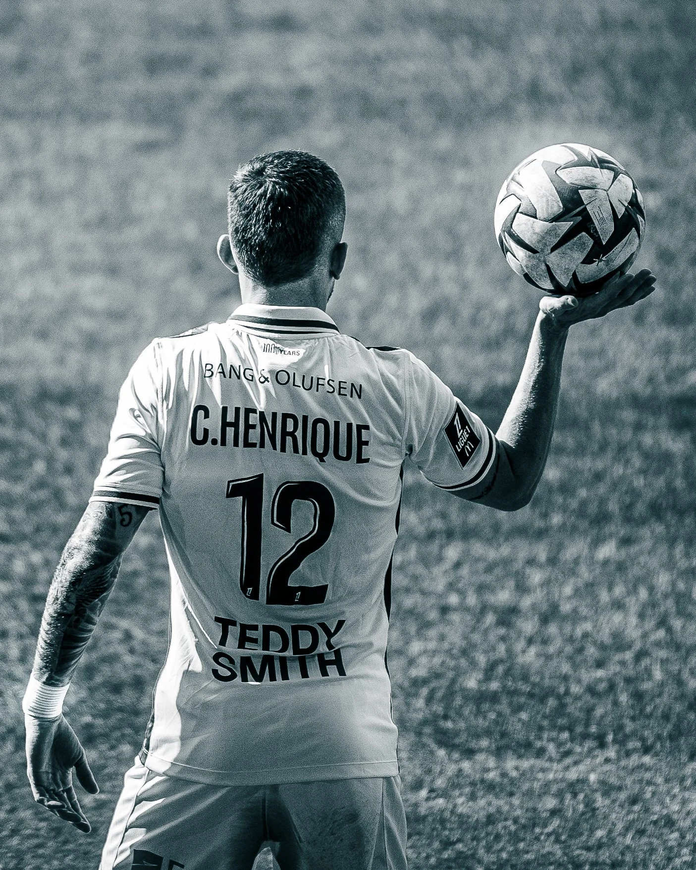 Un footballeur de dos portant un maillot blanc avec le nom "C. HENRIQUE" et le numéro 12. Il tient un ballon de football dans sa main droite.