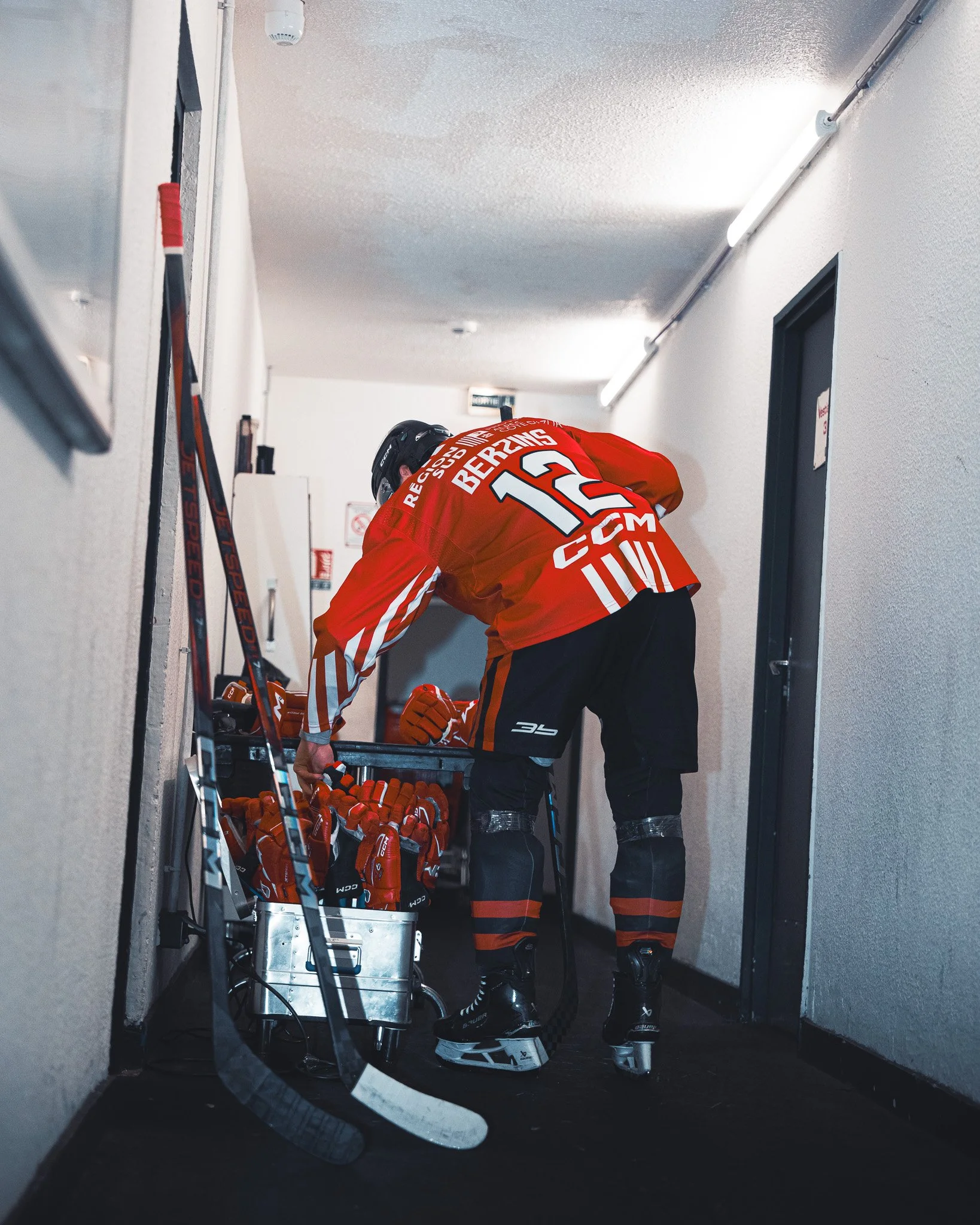 Un joueur de hockey sur glace dans un couloir, portant un maillot rouge avec le numéro 12, et ramassant des gants rouges près de crosses de hockey.