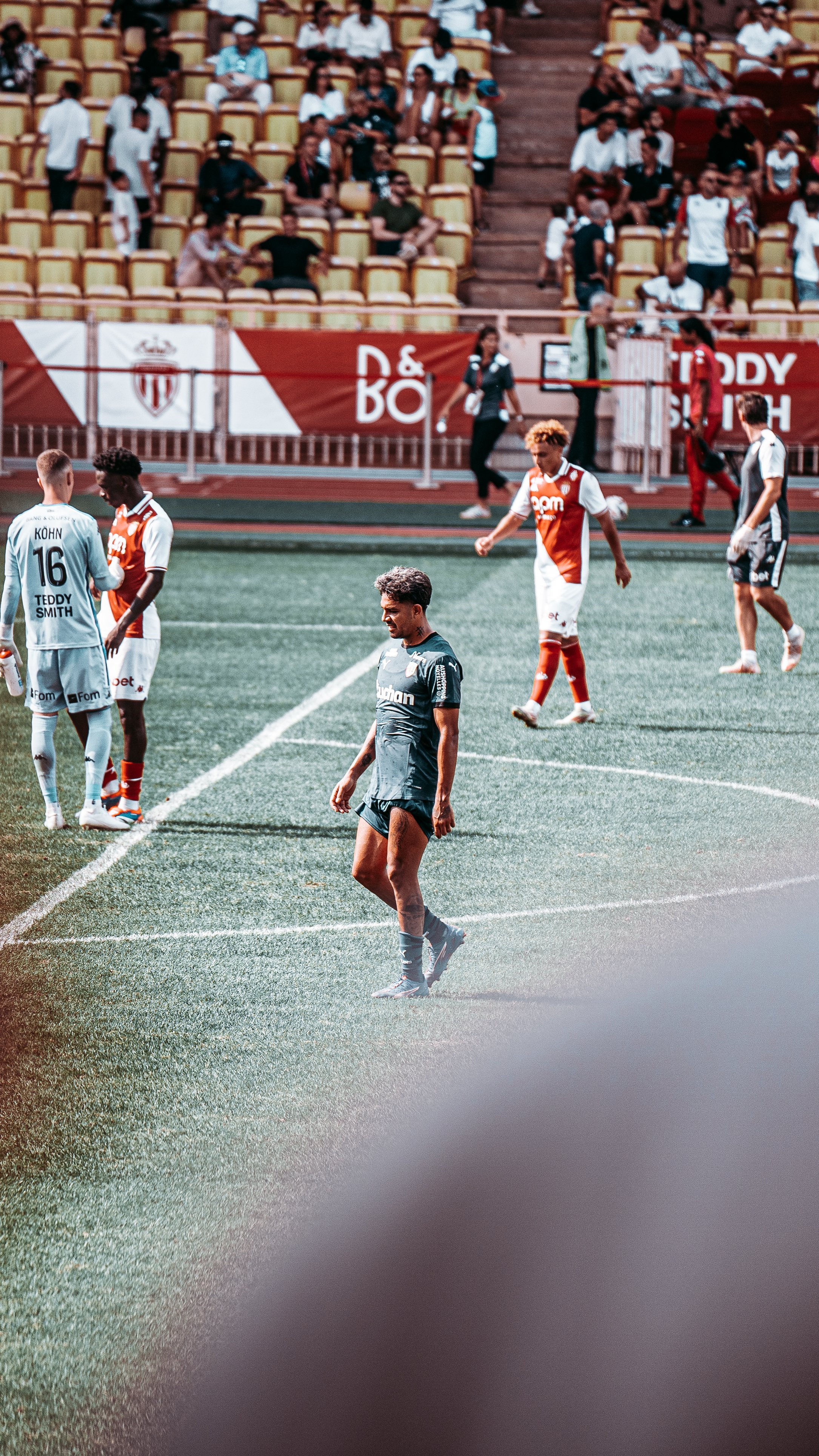 Match de football avec des joueurs en tenue rouge et blanche sur un terrain vert, spectateurs en arrière-plan.