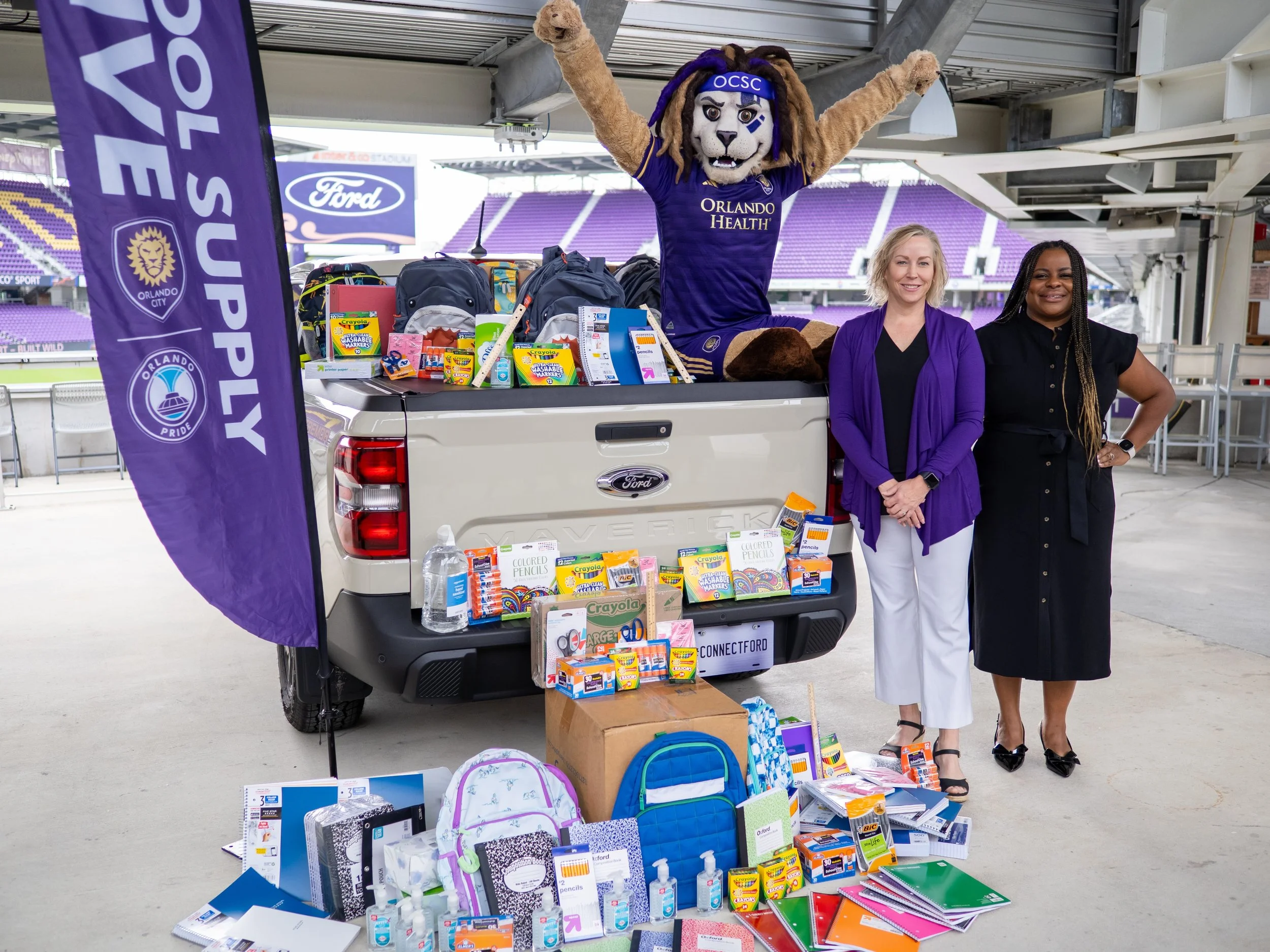 piles of donated school supplies in the back of a Ford pickup truck with Kingston the mascot and two women standing next to him