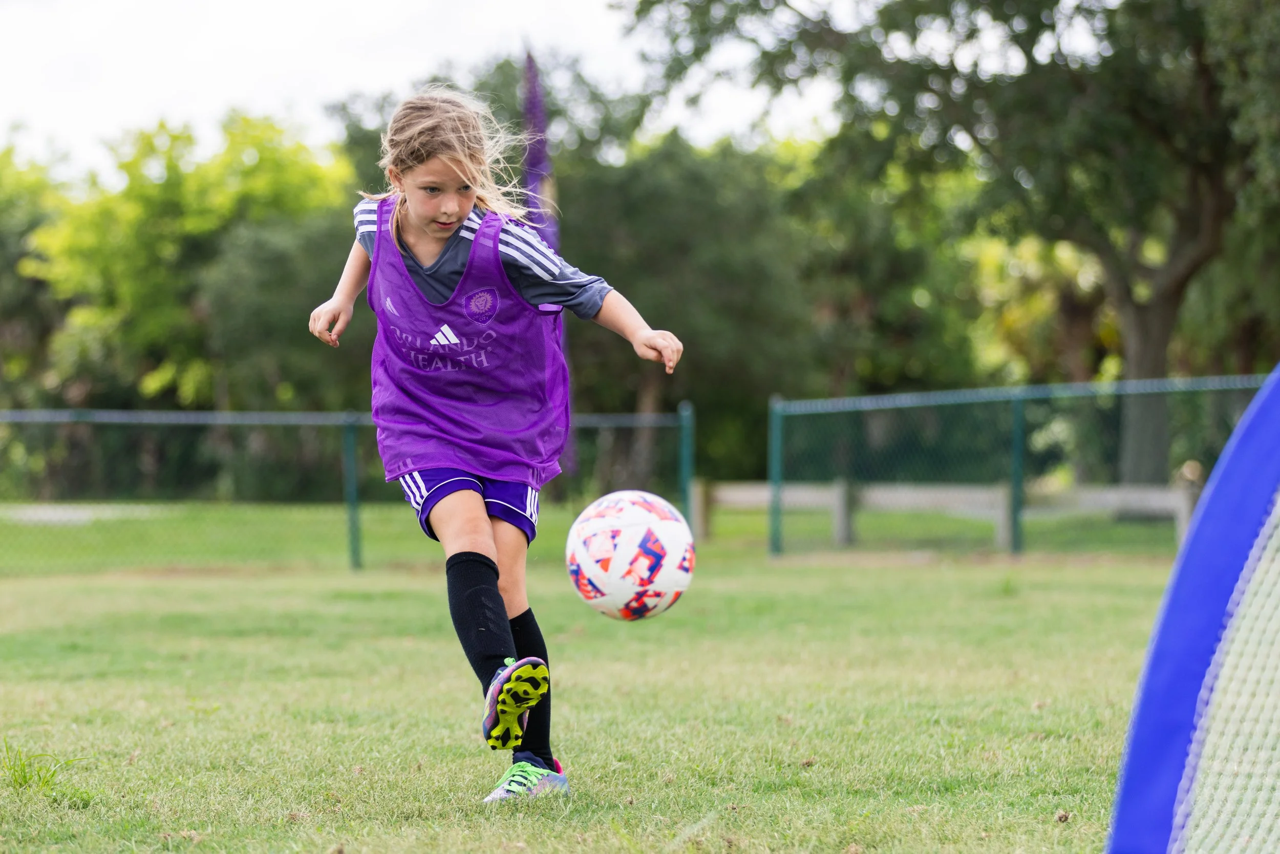 young girl with a purple pinnie on with black socks kicking a soccer ball into a small goal