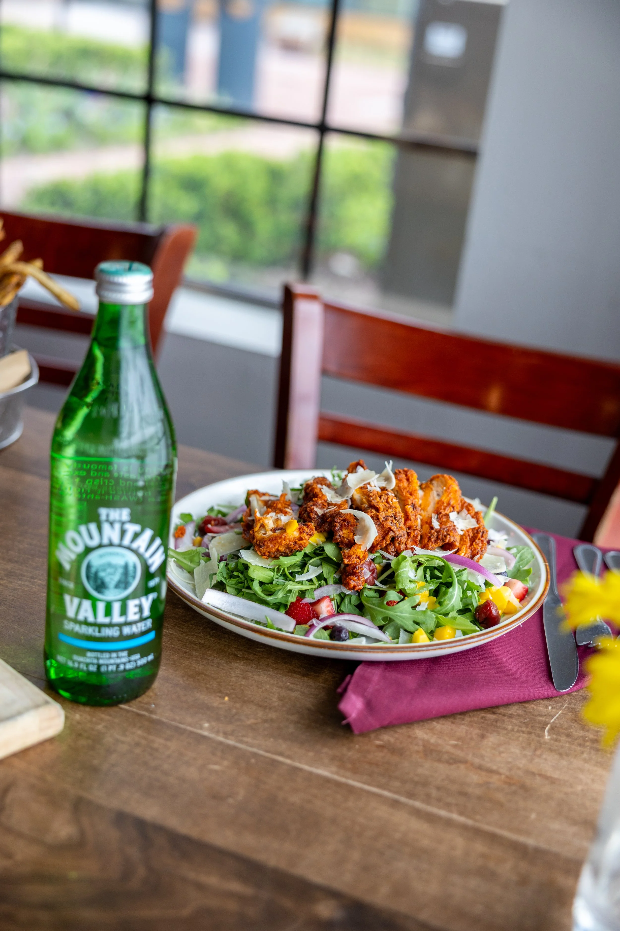 A plate of grilled chicken with tomatoes, boiled eggs, blue cheese, bacon, and greens, with a small cup of dressing on the side, a slice of pizza with arugula, and a glass of champagne on a wooden table.