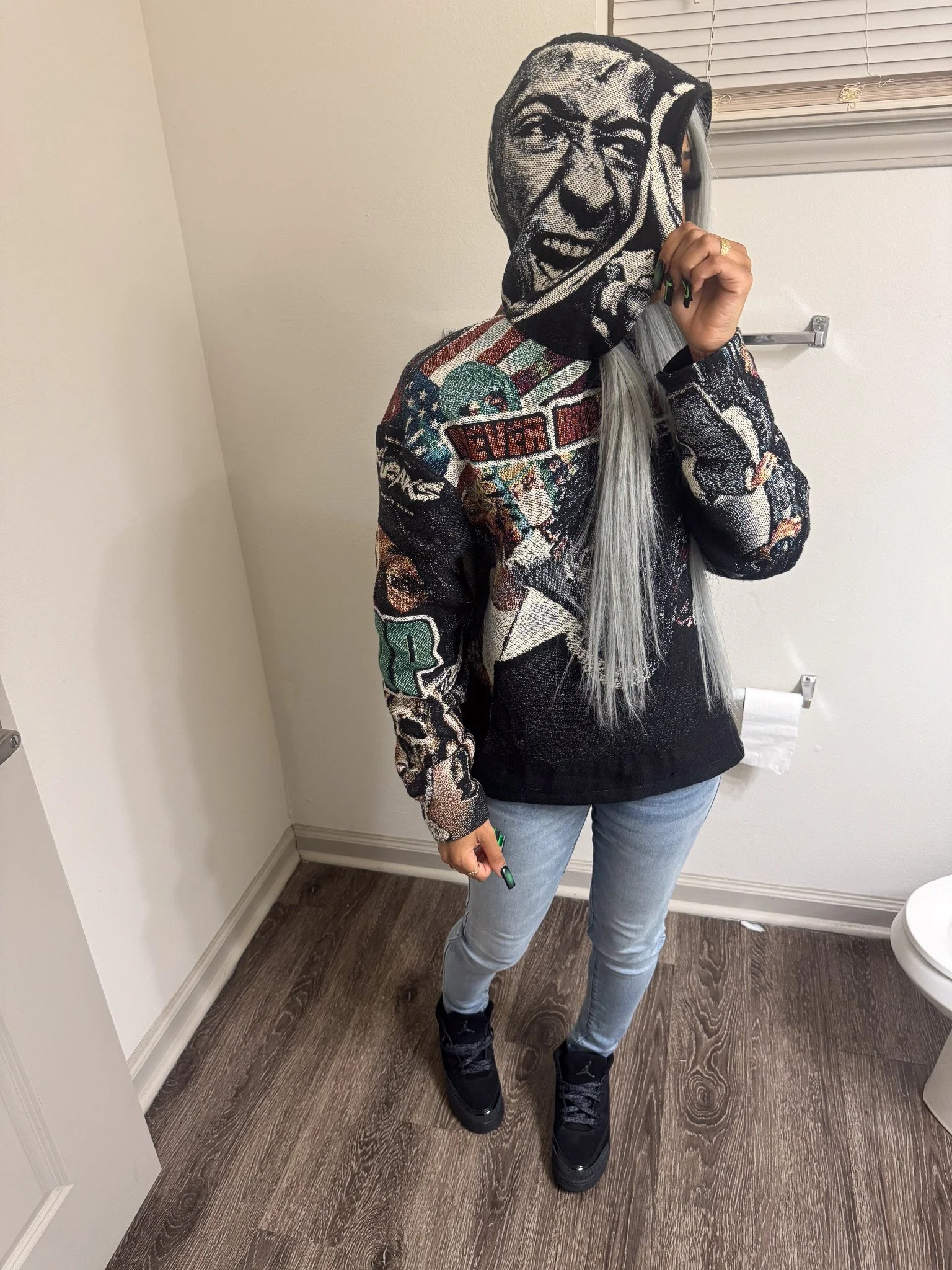 Person wearing a face mask with a large printed face, jeans, and black sneakers, standing in a room with wood flooring and white walls.