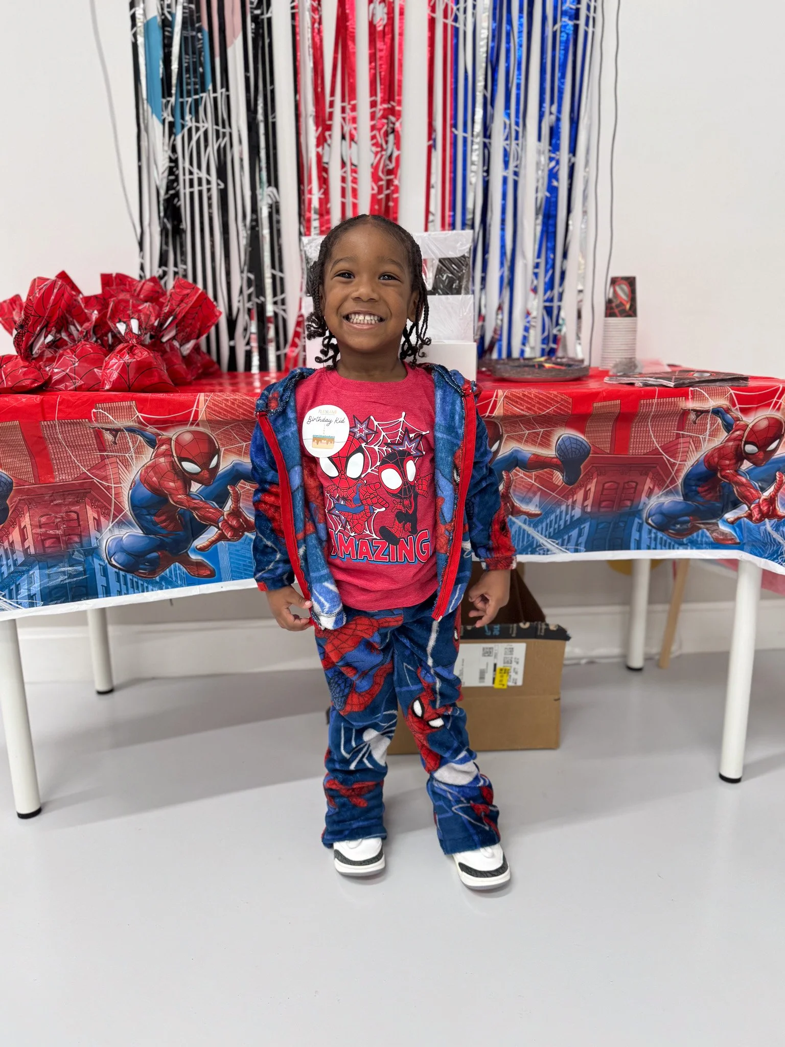 Young boy smiling at a Spider-Man themed birthday party, standing in front of a table with Spider-Man decorations and party favors.