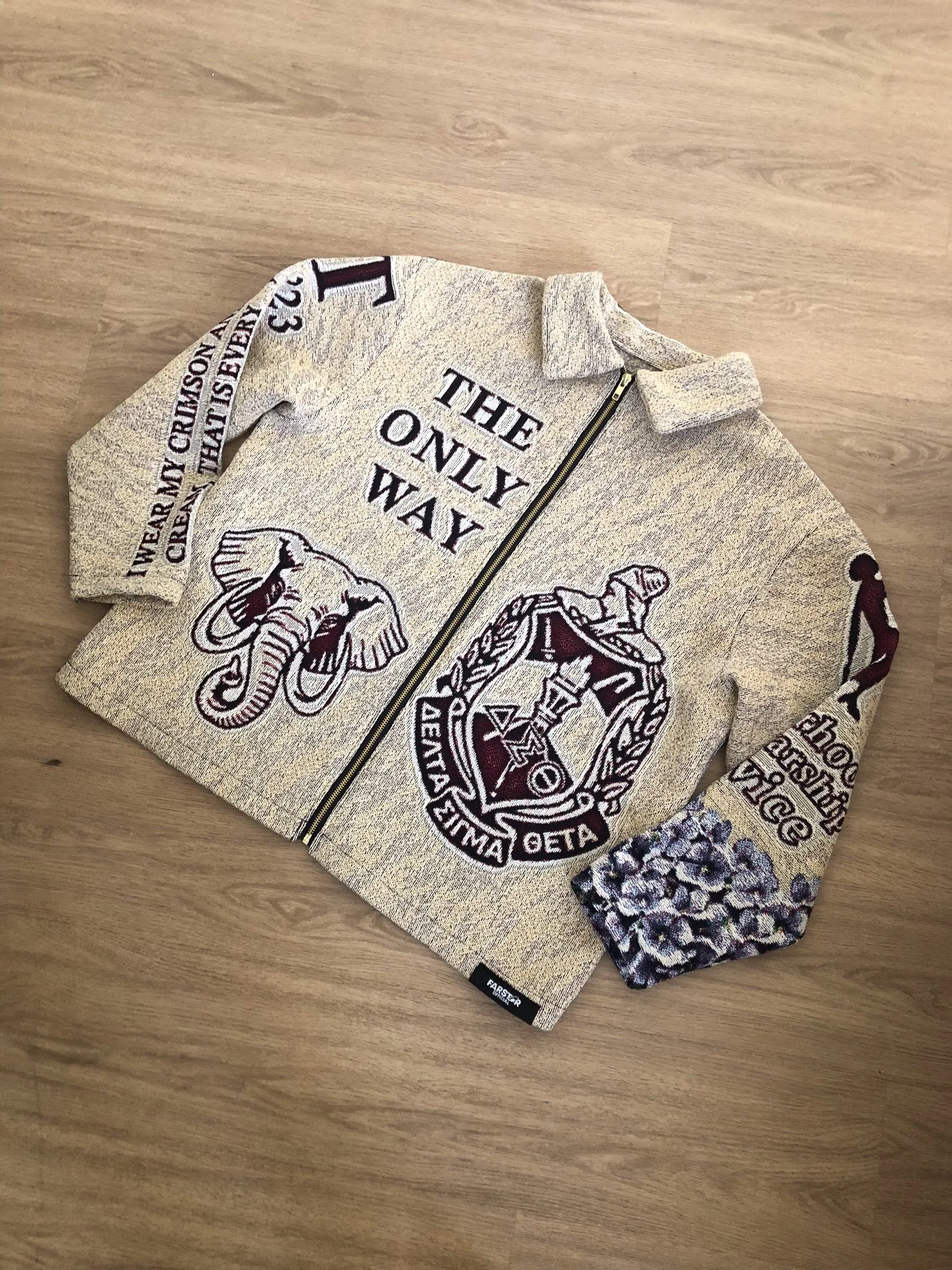 A beige hoodie with military patches, a skull design, and various patches and lettering on the sleeves, lying on a wooden floor.