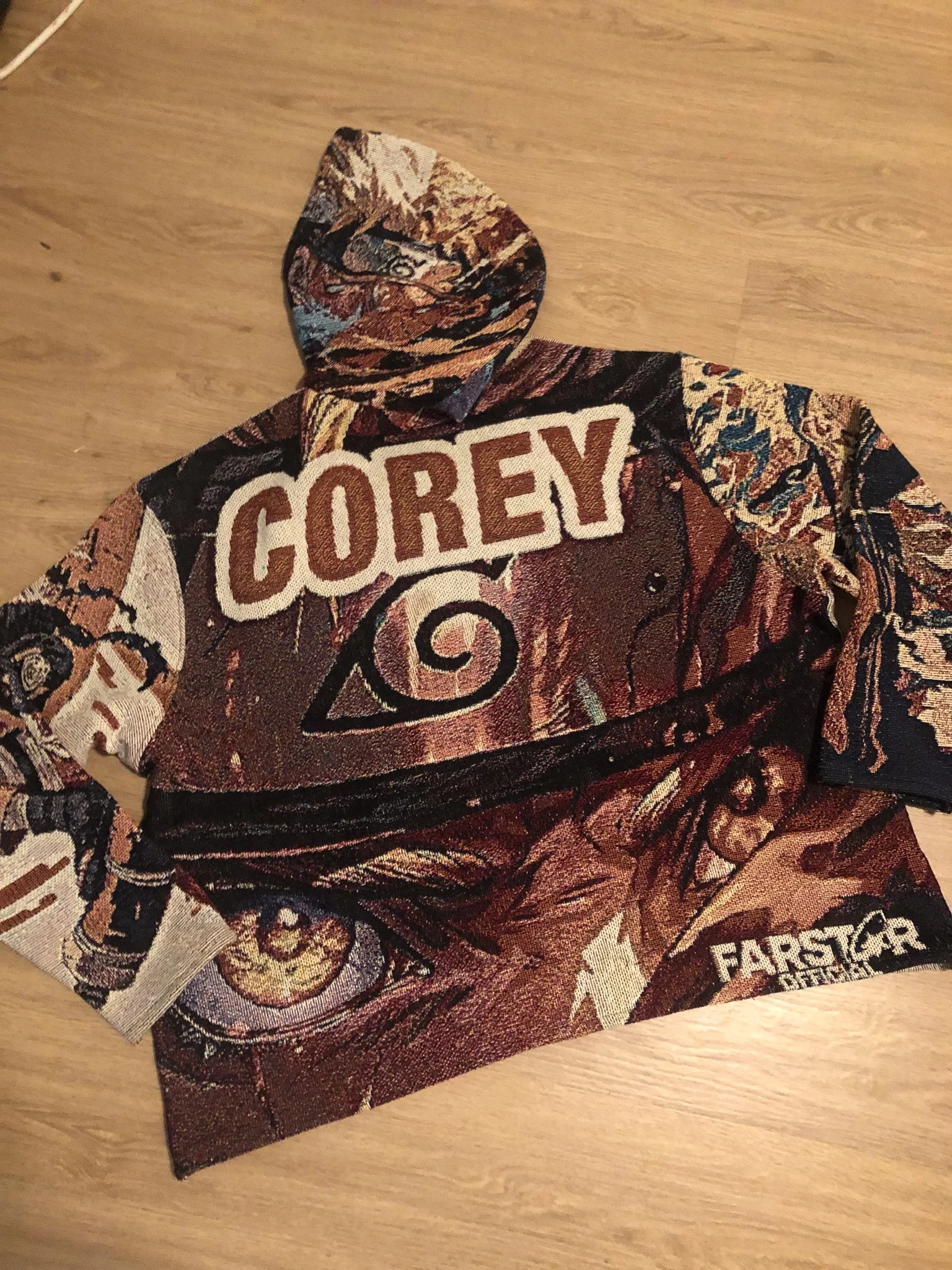 Close-up of a fabric item, possibly a piece of clothing or a towel, with a colorful, intricate pattern and a large embroidered patch that reads 'COREY.' The fabric features various images, including a skull with flowers and other decorative elements.